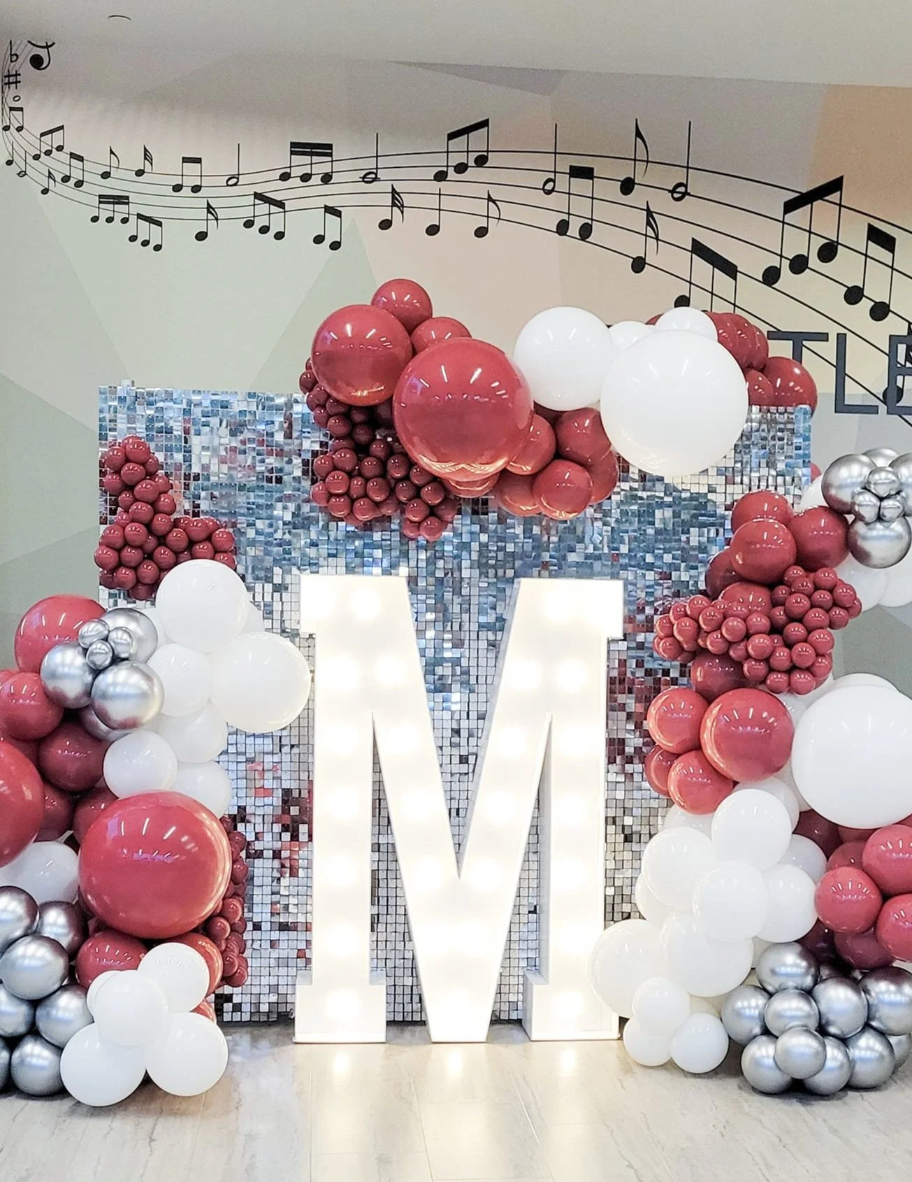 Decorative display with a large illuminated letter 'W' surrounded by clusters of pink, white, and silver balloons, set against a backdrop of a mosaic mirror and music notes on the wall.