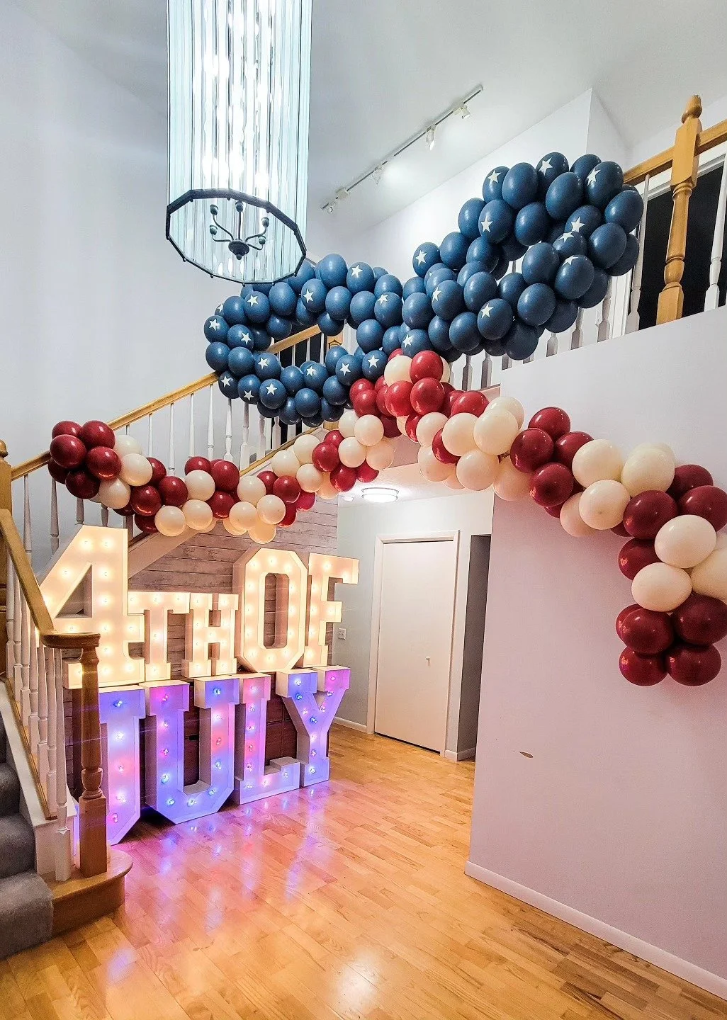 Decorative interior with a balloon arch in red, white, and blue, large illuminated '4TH OF JULY' sign, and a modern chandelier hanging from the ceiling.
