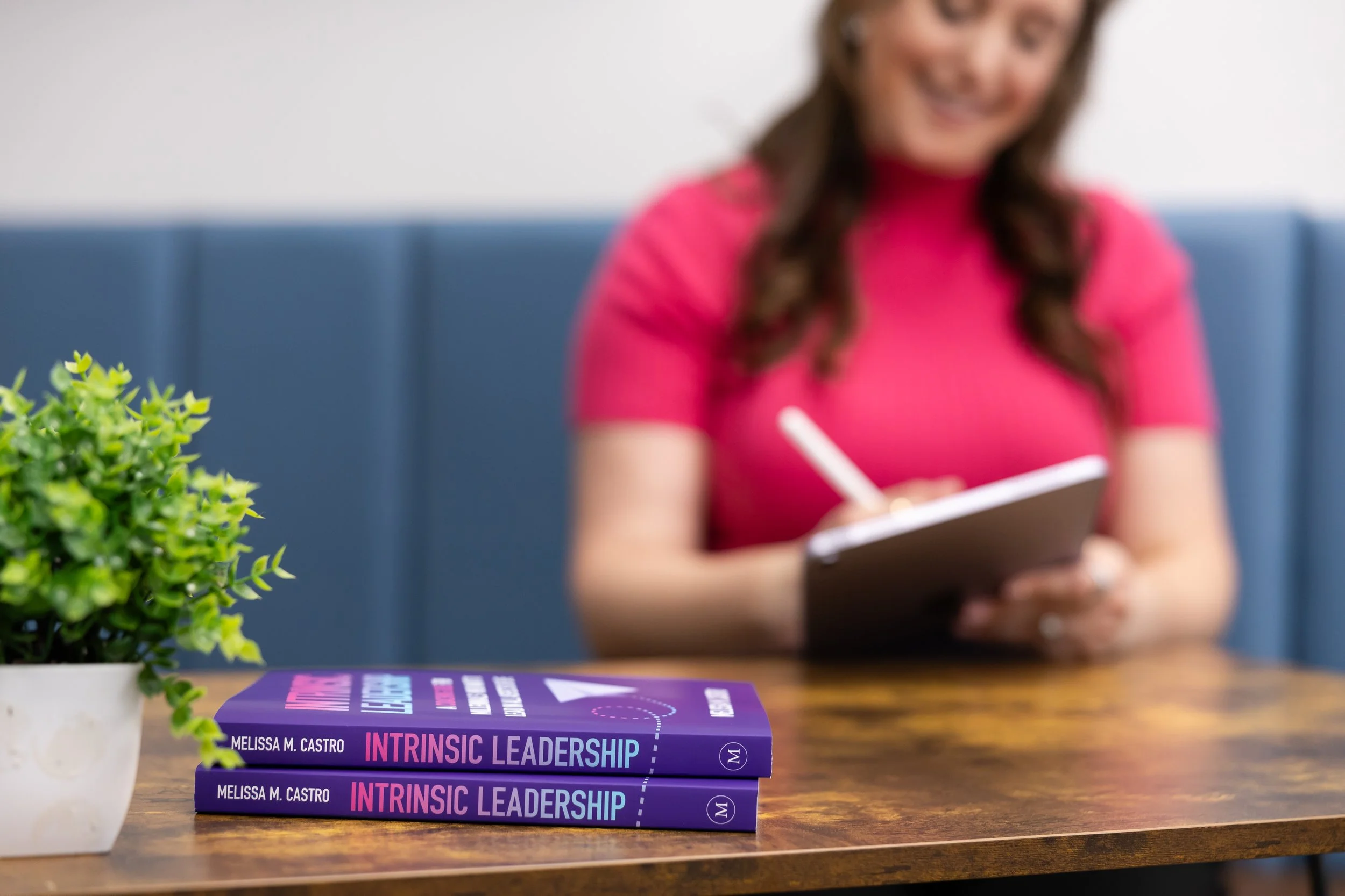 A table with a green potted plant and two copies of the book 'Intrinsic Leadership' by Melissa M. Castro, with a woman in a pink shirt writing in a notebook in the background.