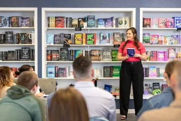 A woman giving a talk or reading in front of a bookshelf while an audience of young adults listens.