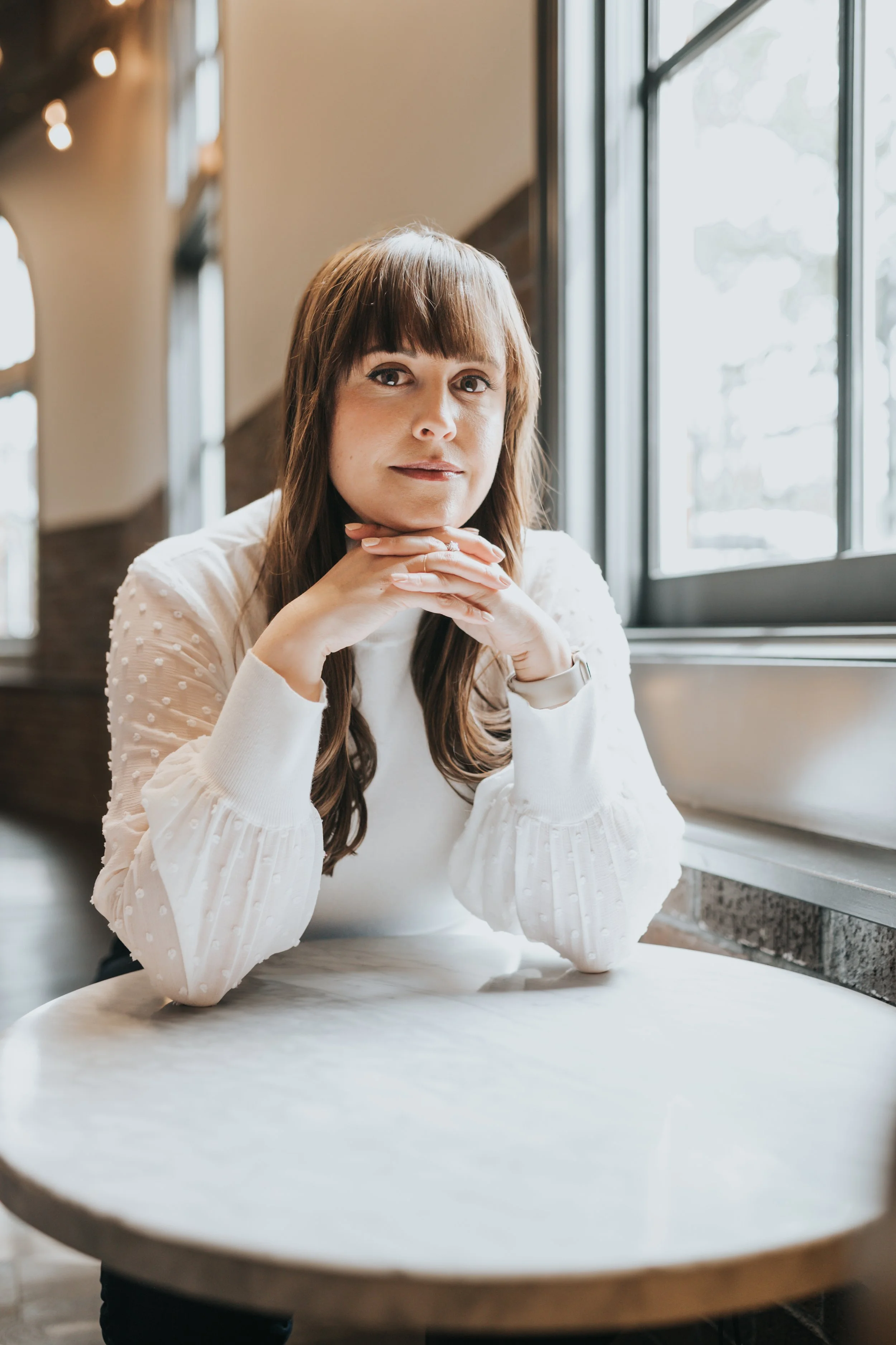 A woman with long brown hair and bangs, wearing a white blouse with textured dots, sits at a table near a large window in what appears to be a cafe or restaurant, resting her chin on her hands and looking at the camera.