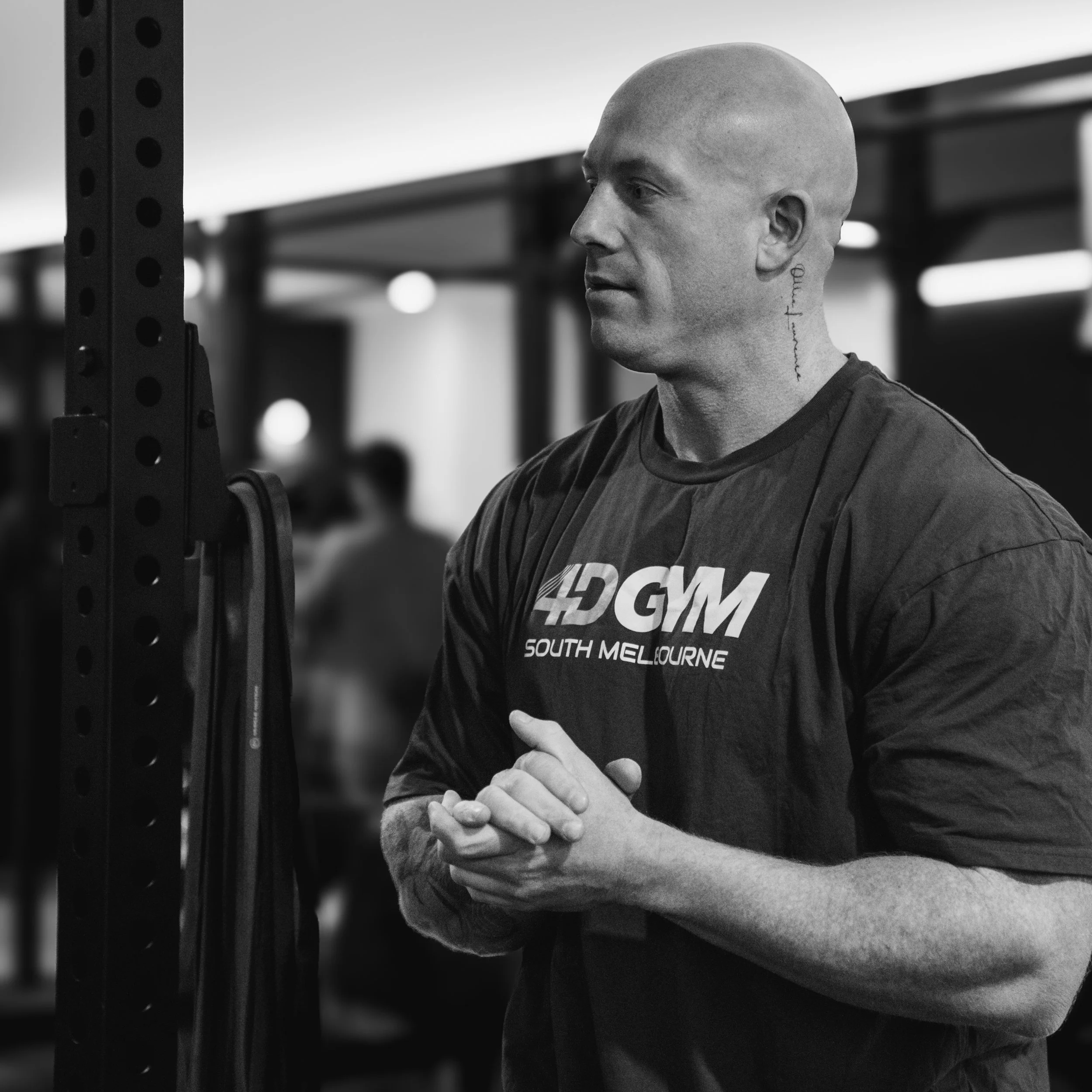 A bald man with tattoos on his neck is standing in a gym, wearing a black t-shirt with the logo '4D GYM South Melbourne'. He appears to be paying attention to something or someone off-camera. The background is blurred, showing gym equipment and other people working out.