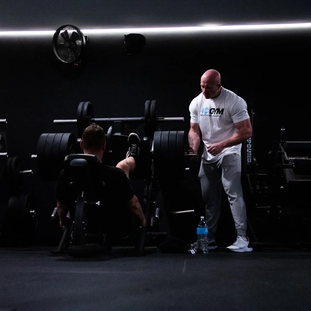 A man in a black shirt sits on a workout machine with his legs extended, while a bald man in a white t-shirt and gray sweatpants stands next to him inside a gym with black walls and various weightlifting equipment.