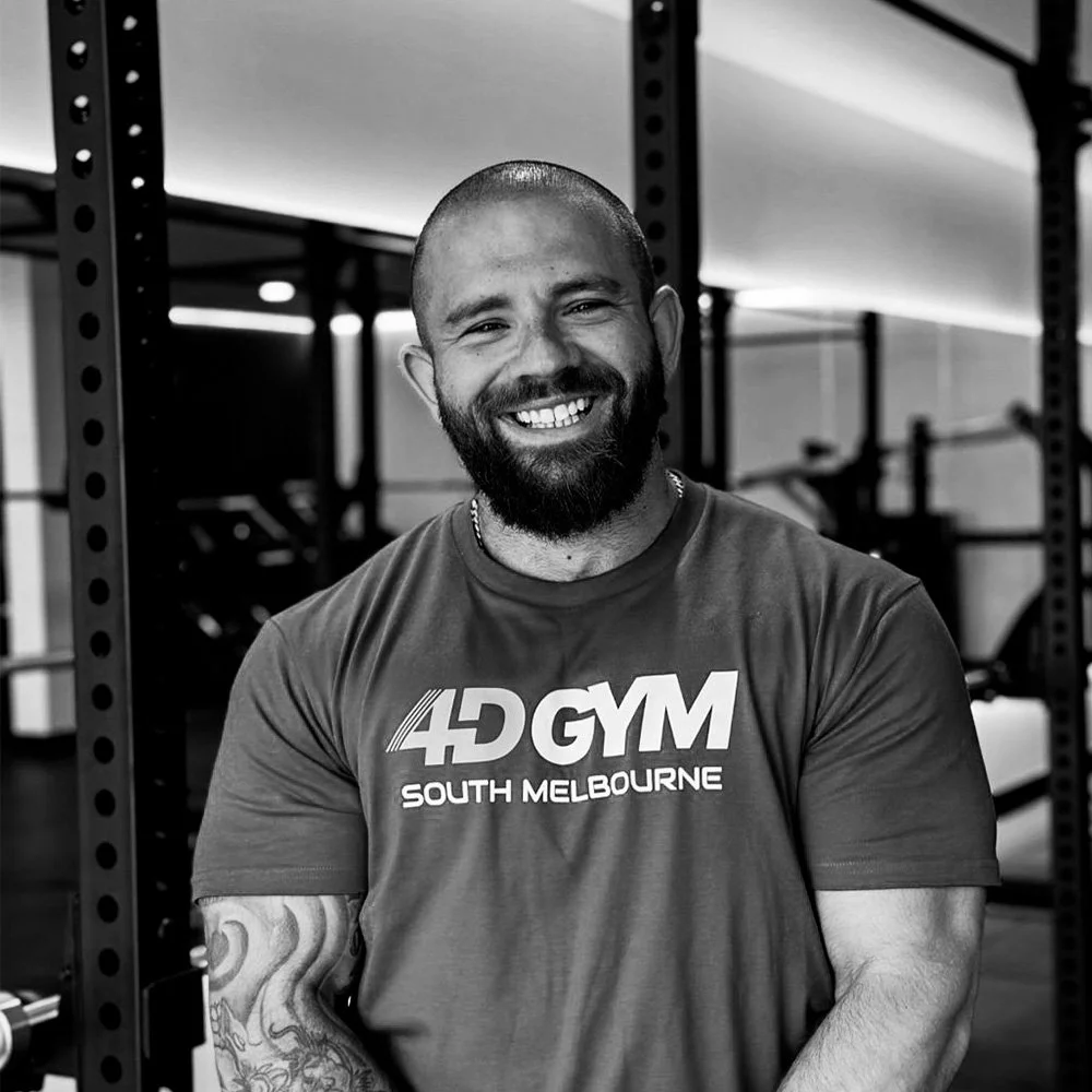 A smiling man with a beard and tattoo wearing a gym shirt labeled 4D GYM South Melbourne in a gym with weightlifting equipment.