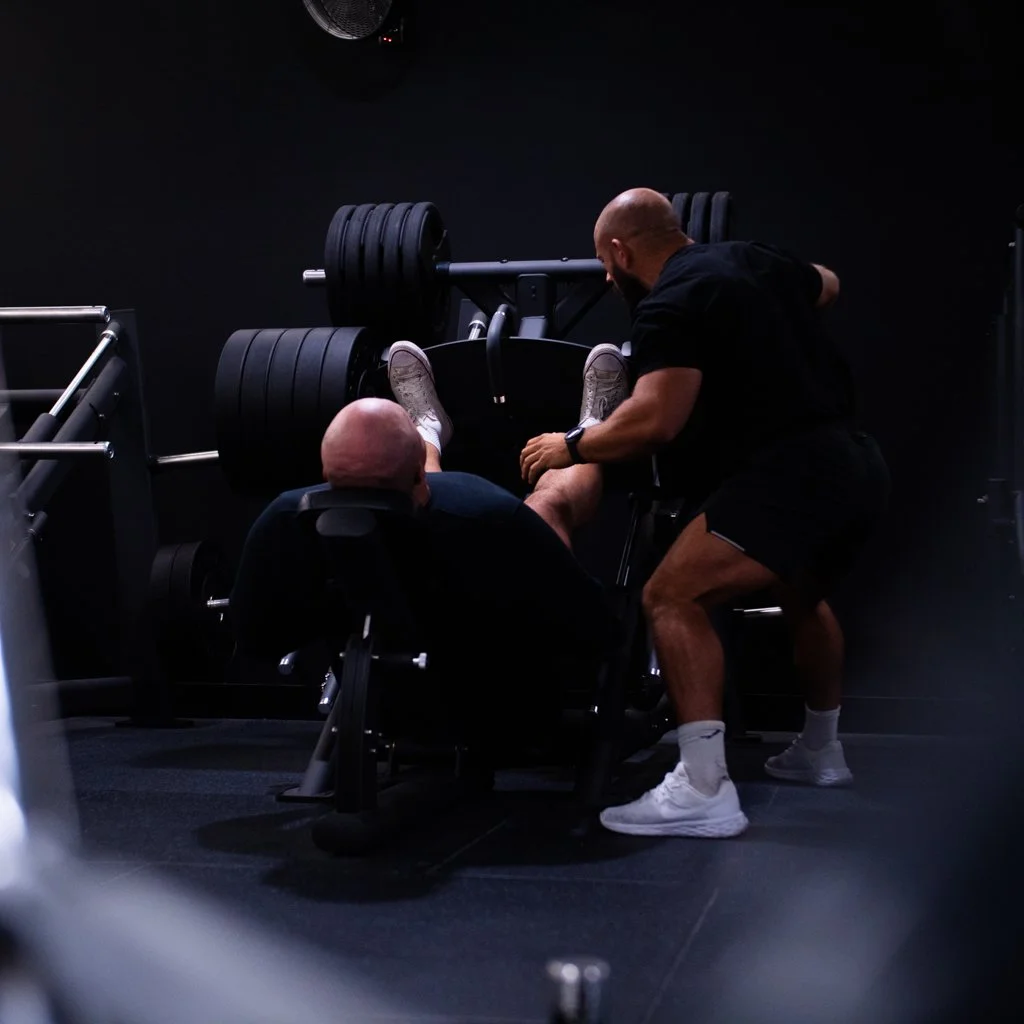 A man being assisted with a leg press exercise at the gym by a trainer.