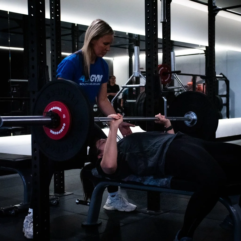 A woman spotting another woman doing a bench press at the gym.