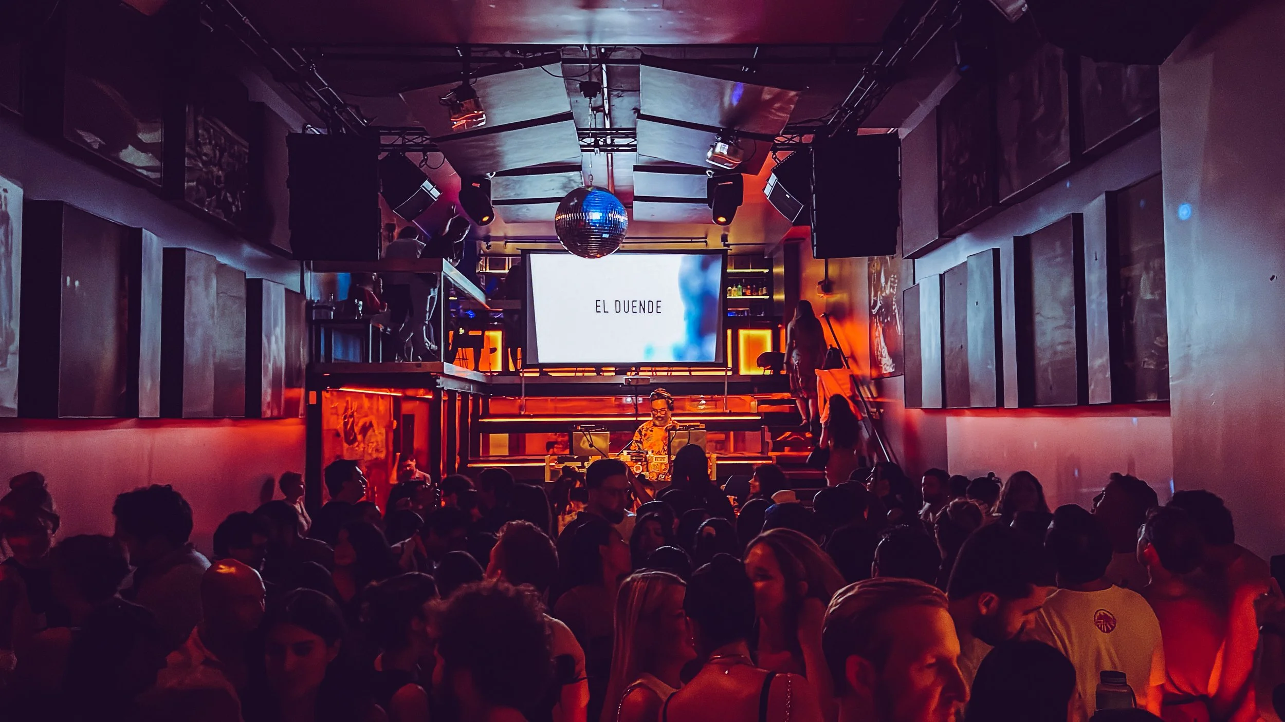 A lively nightclub scene with a DJ performing on stage in front of a large screen displaying 'EL DUENDE'. The dance floor is crowded with people dancing and socializing. The room has vibrant red and purple lighting, with a disco ball hanging from the