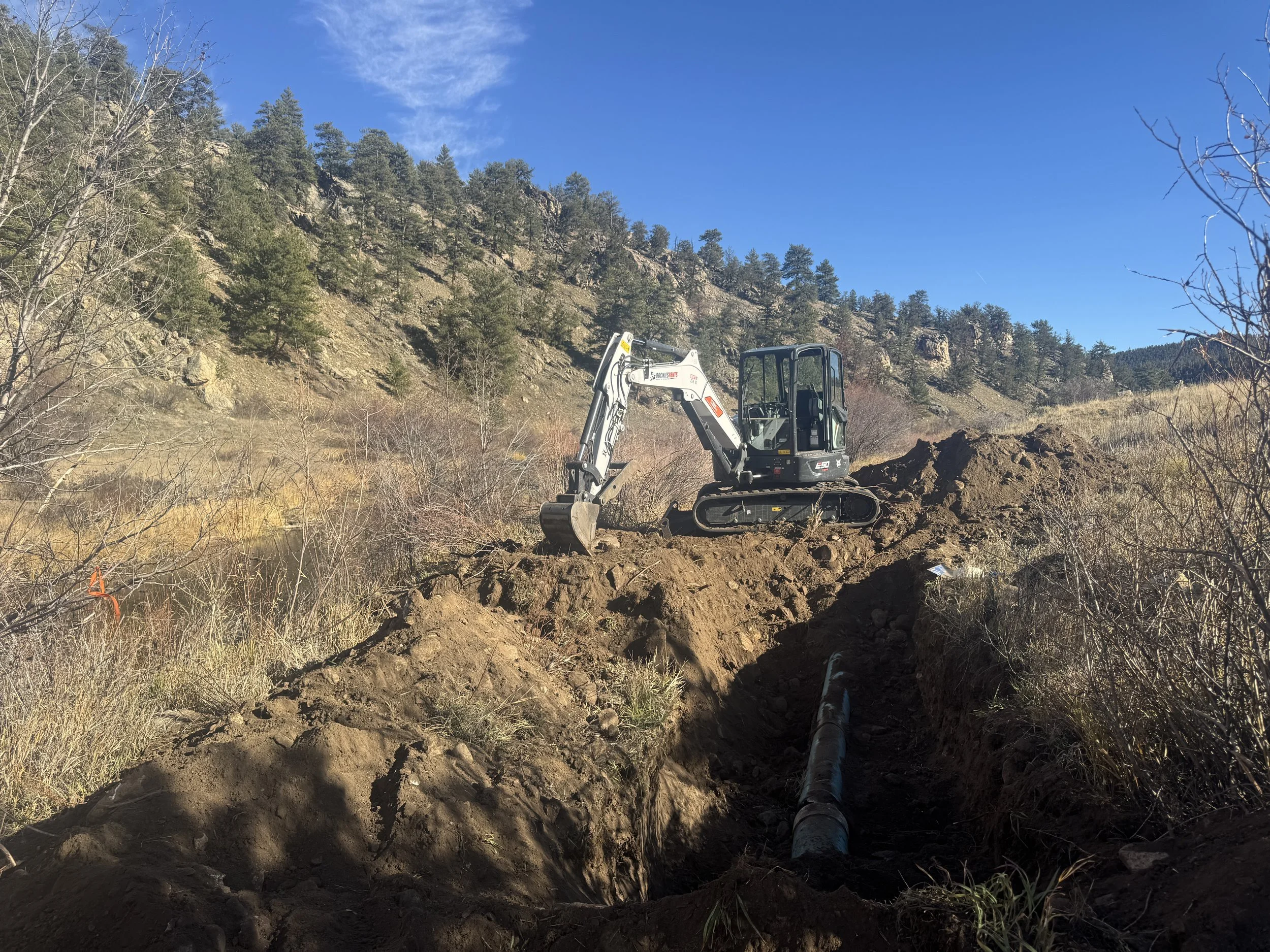 A small excavator working on a construction site in a hilly, rural area with dirt, sparse trees, and a blue sky.