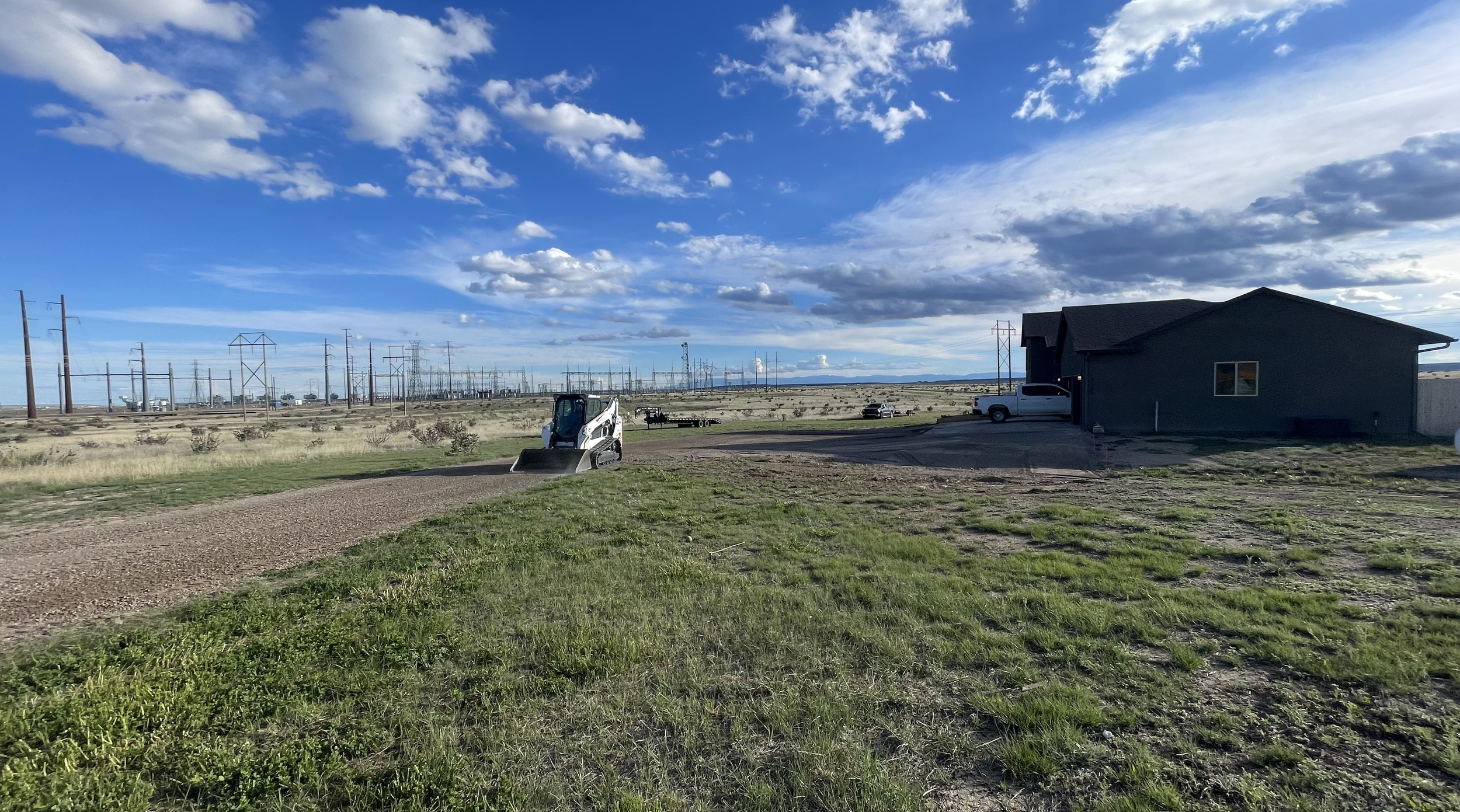 A landscape with a gravel driveway leading to a gray house, a small white vehicle with a scraper attachment, a pickup truck, and a horse trailer. In the background, there are power lines and an industrial area under a partly cloudy sky.