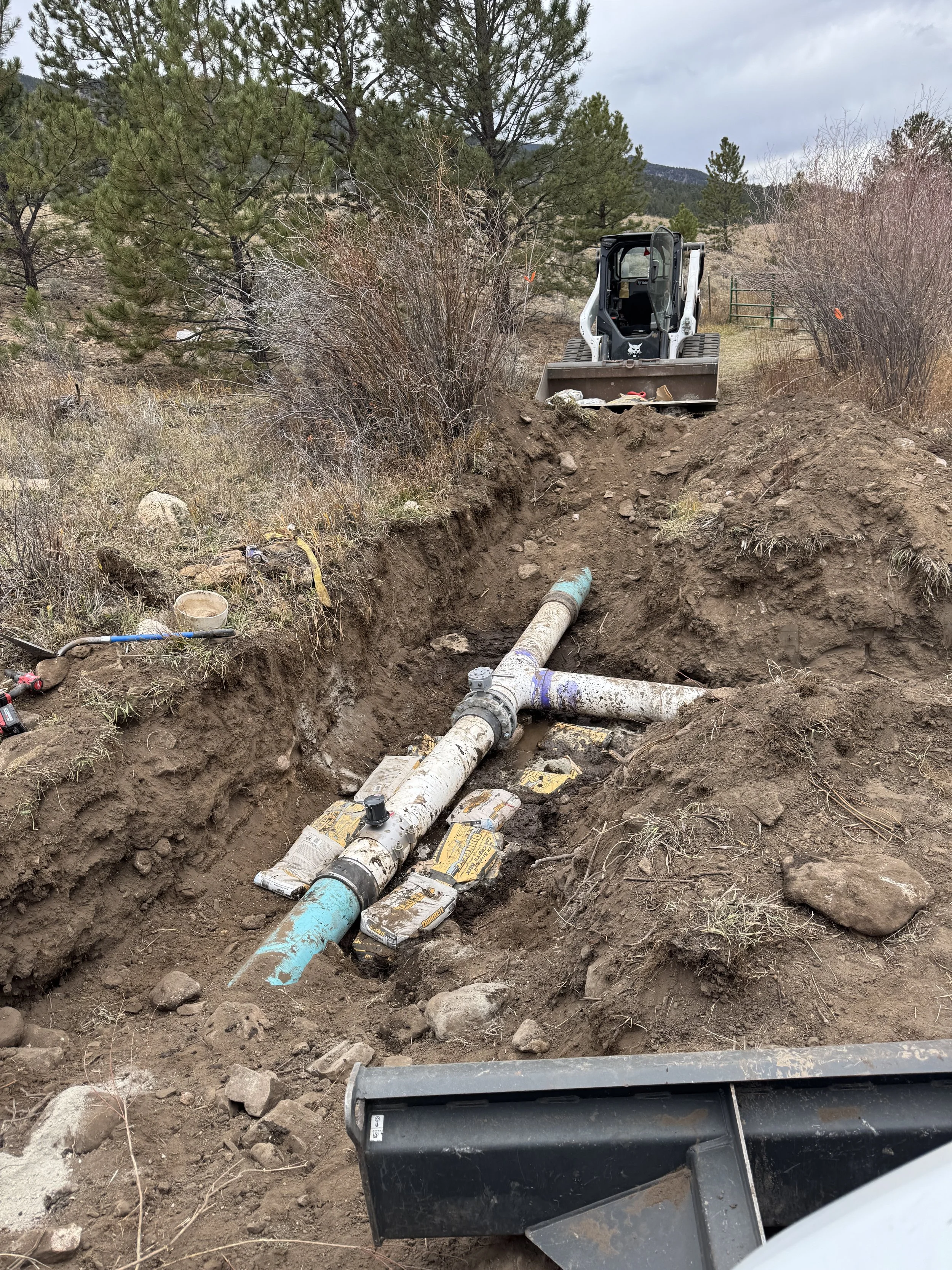 Construction site with a trench for installing or repairing water or sewer pipes, a small excavator at the end of the trench, and a clear sky in the background.