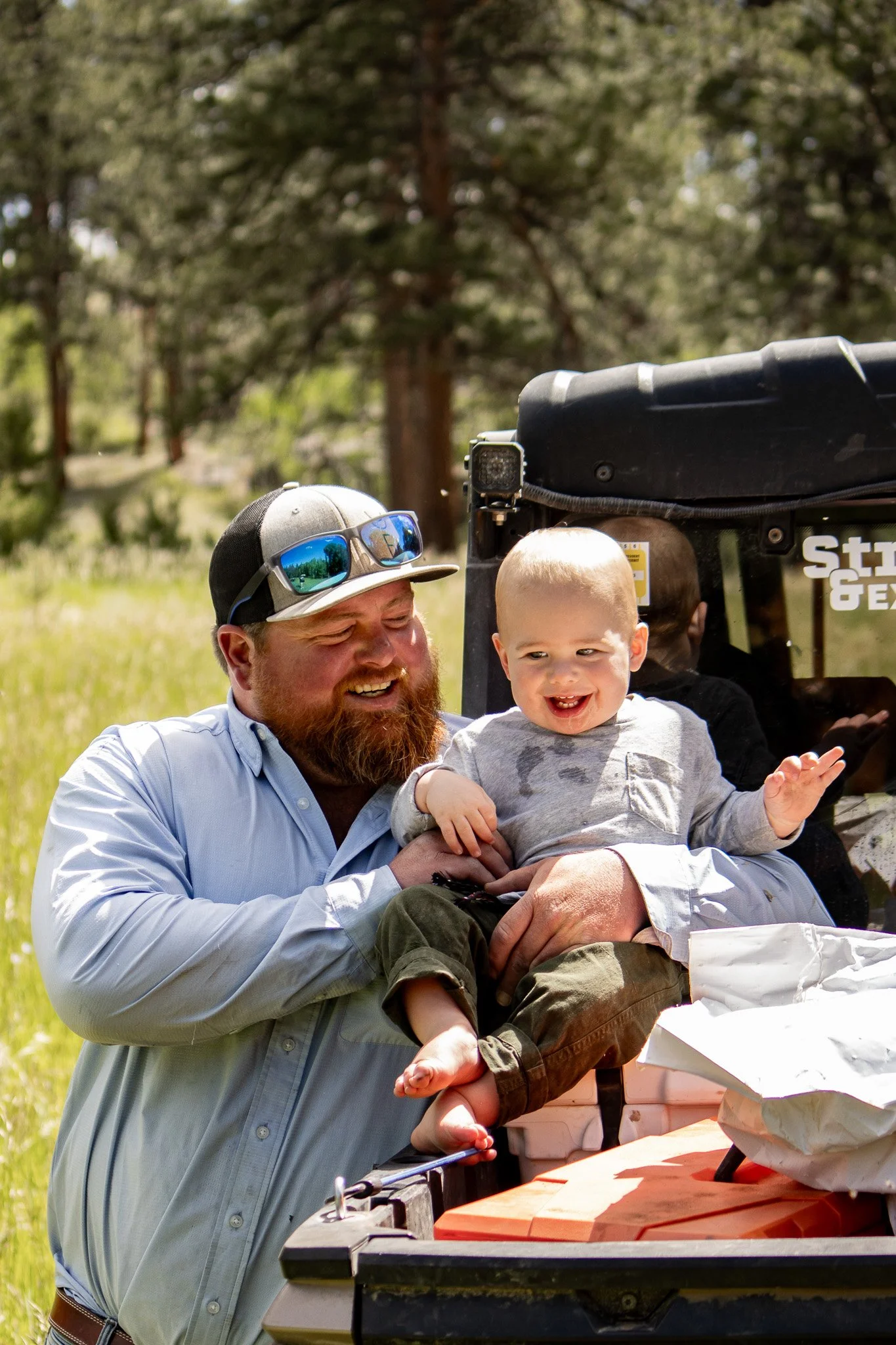 A man with sunglasses on his cap holding a smiling young boy on a vehicle outdoors in a grassy area surrounded by trees.