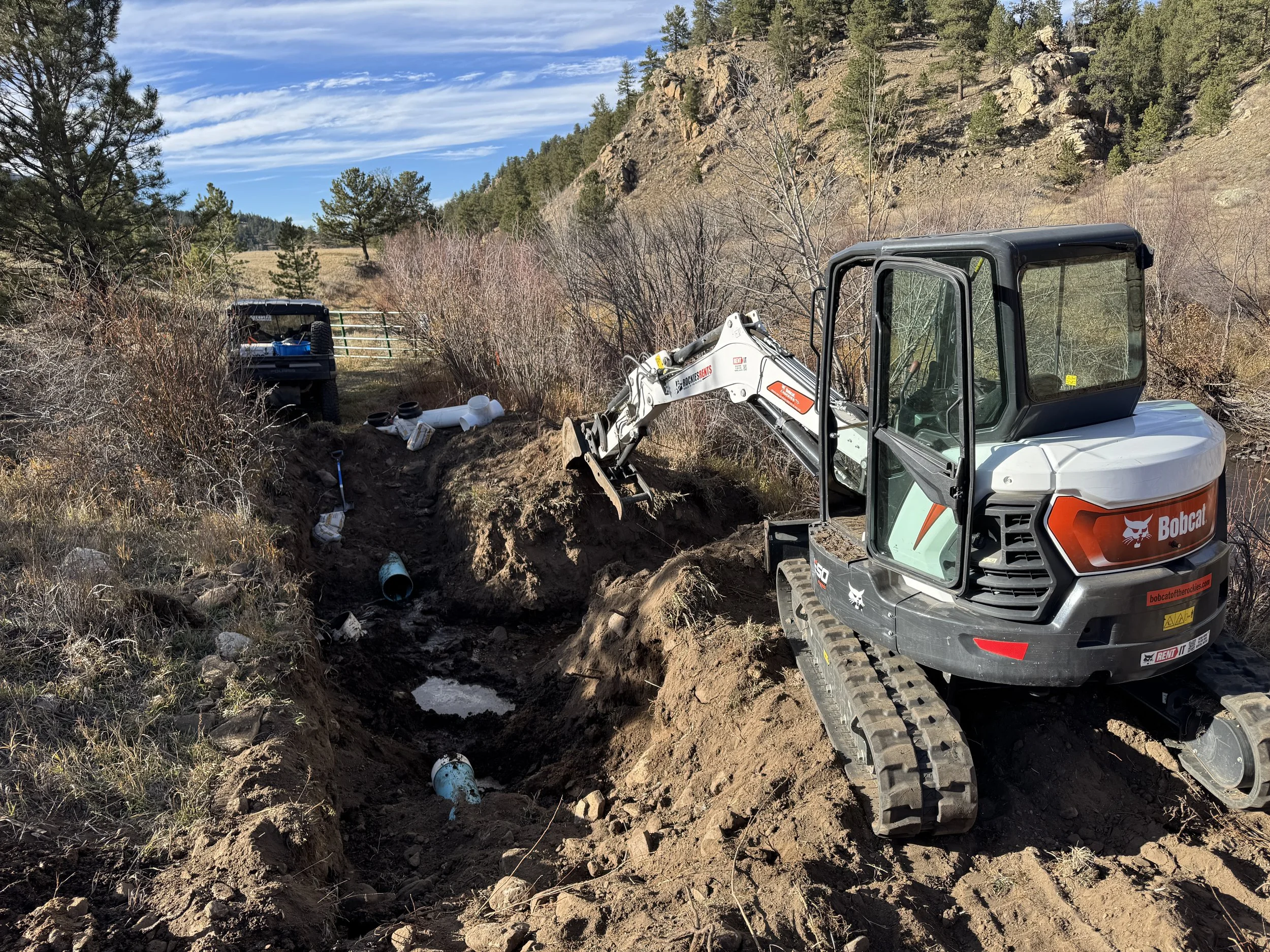 A Bobcat mini excavator is digging a trench in a rural area with a hillside and trees in the background, near a truck with construction tools and pipes.