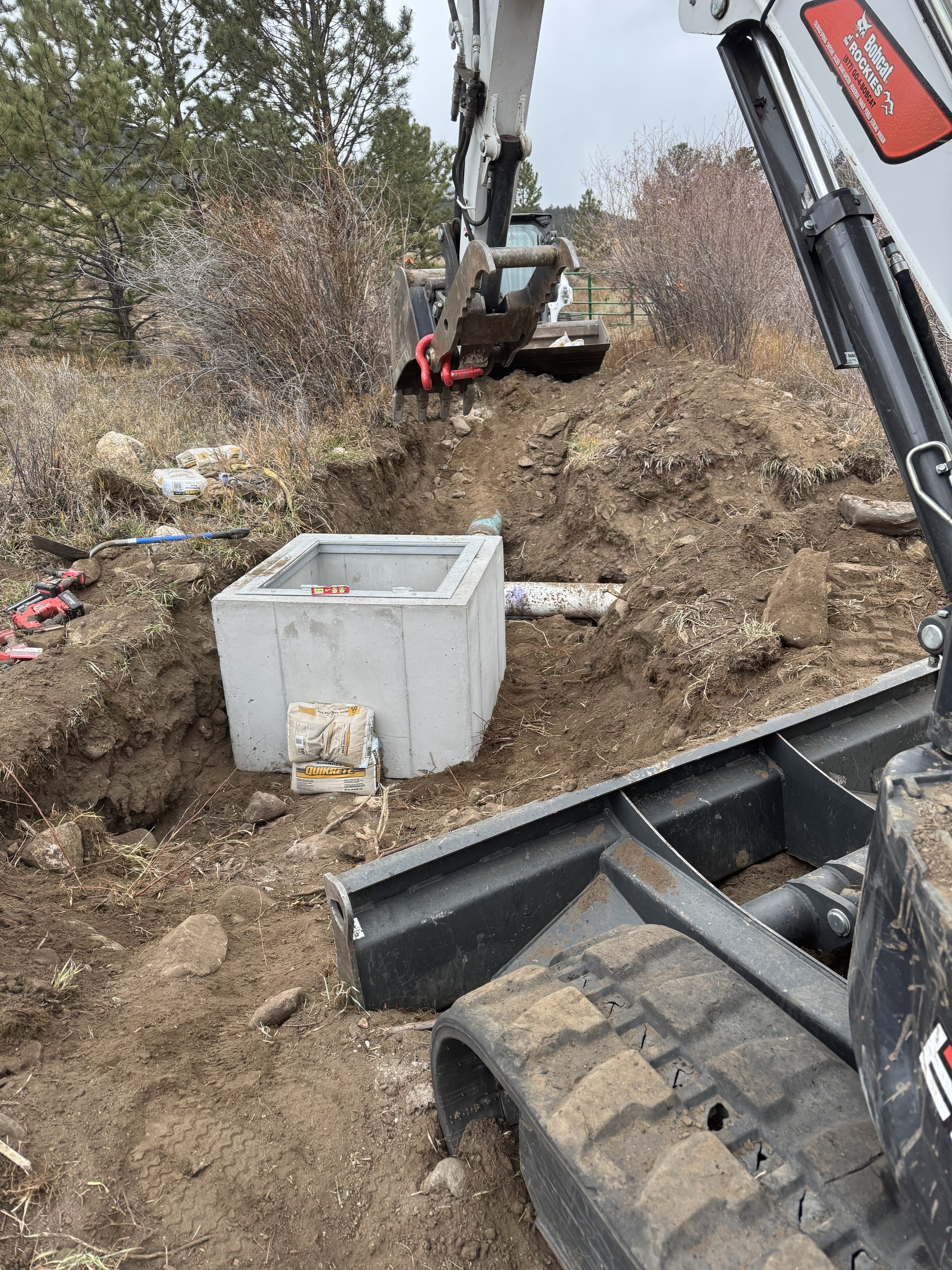 Construction site with excavator installing or repairing underground water or sewer pipes, with concrete box, soil, and tools around.