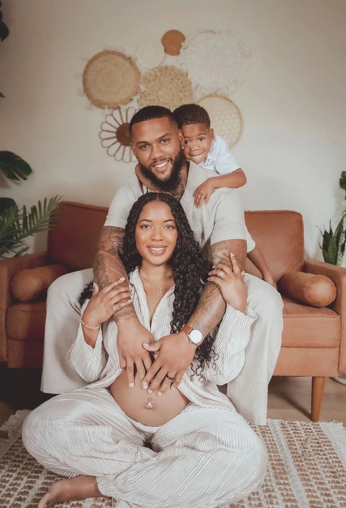 A joyful family portrait with a woman, a man, and a young boy in a cozy indoor setting. The woman is sitting on the floor with her legs crossed, showing her pregnant belly, and the man is sitting behind her with his arms around her. The young boy is playfully on the man's back, leaning on his shoulder. They all have warm smiles, and the background features a wall with decorative weaving art and some plants on either side.