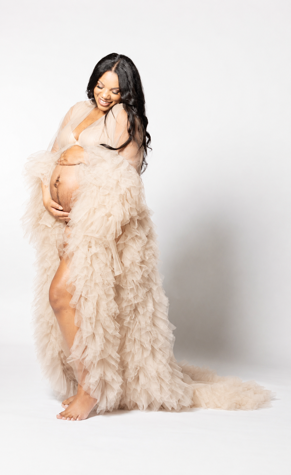A pregnant woman with long black hair smiling, wearing a beige, ruffled gown, holding her belly, standing barefoot against a white background.