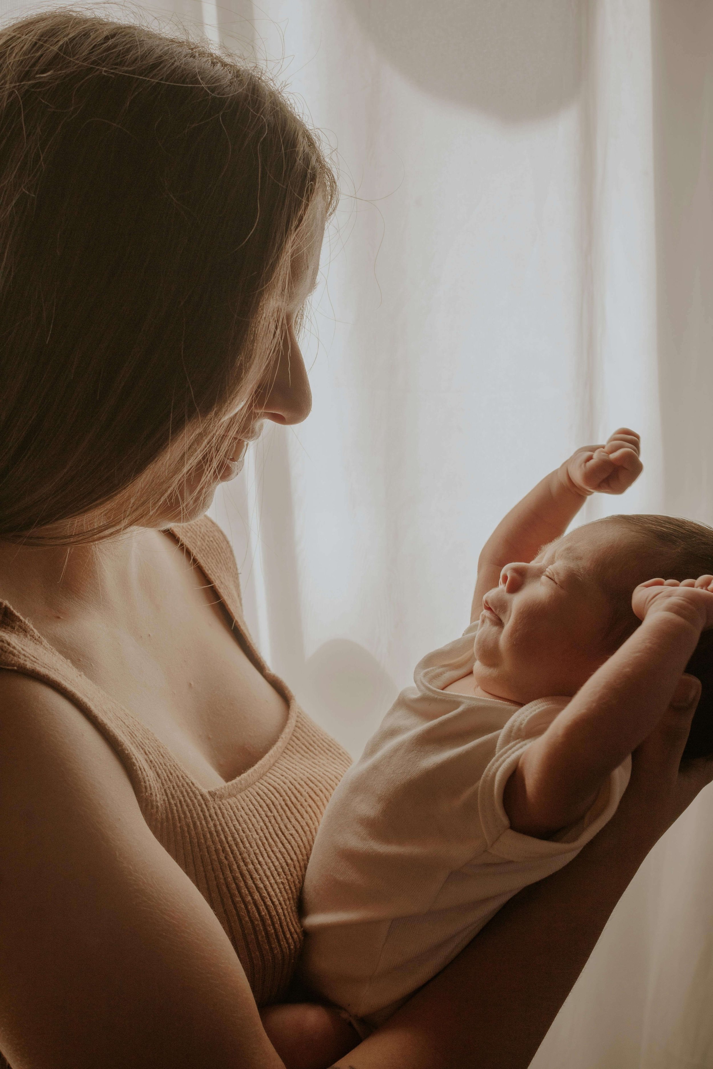 A woman holding a smiling baby, both with light skin, near a light-colored curtain.