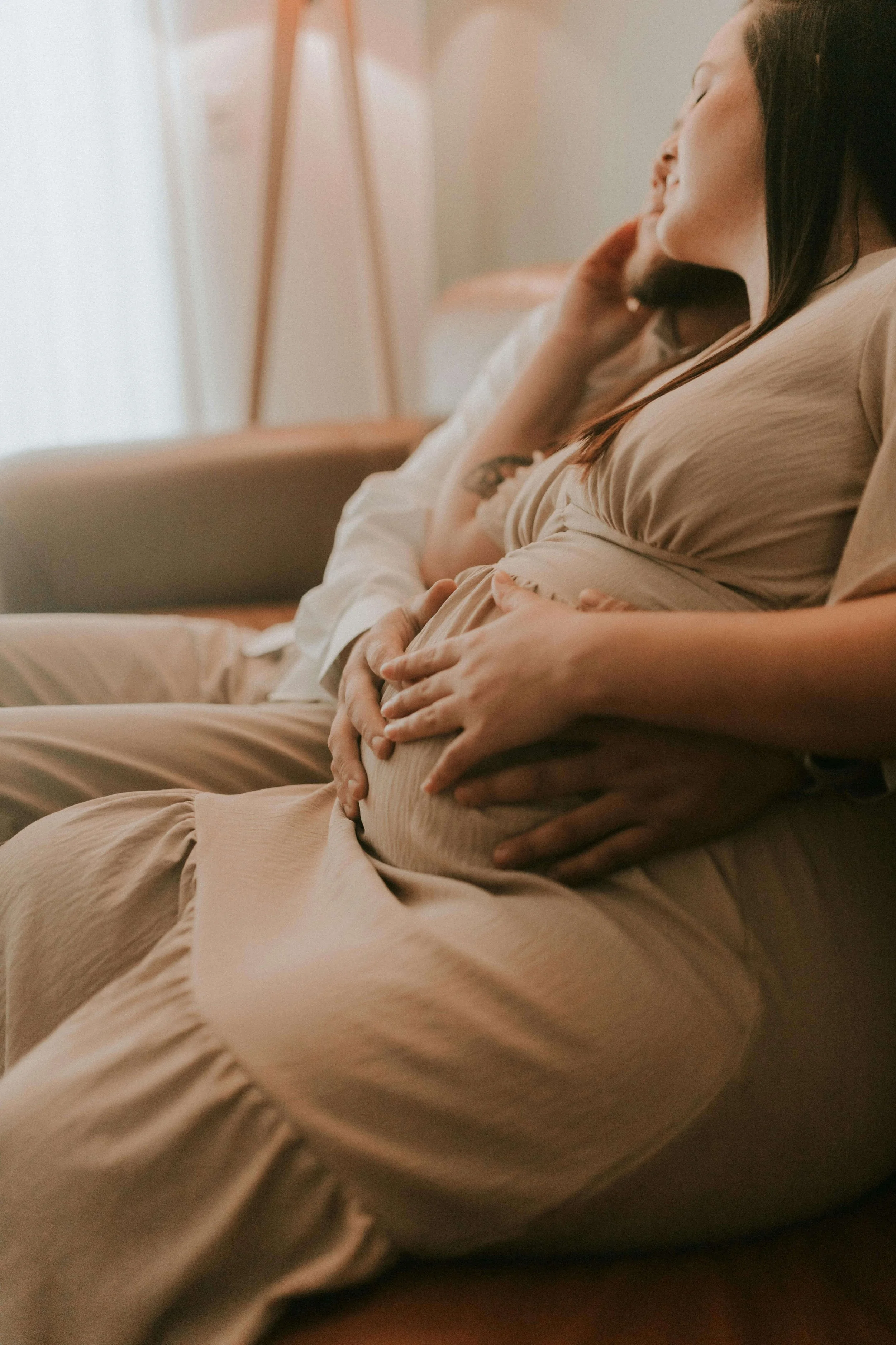 Multiple people sitting on a couch, with one person prominently holding their pregnant belly, relaxing with eyes closed in a cozy, softly lit room.