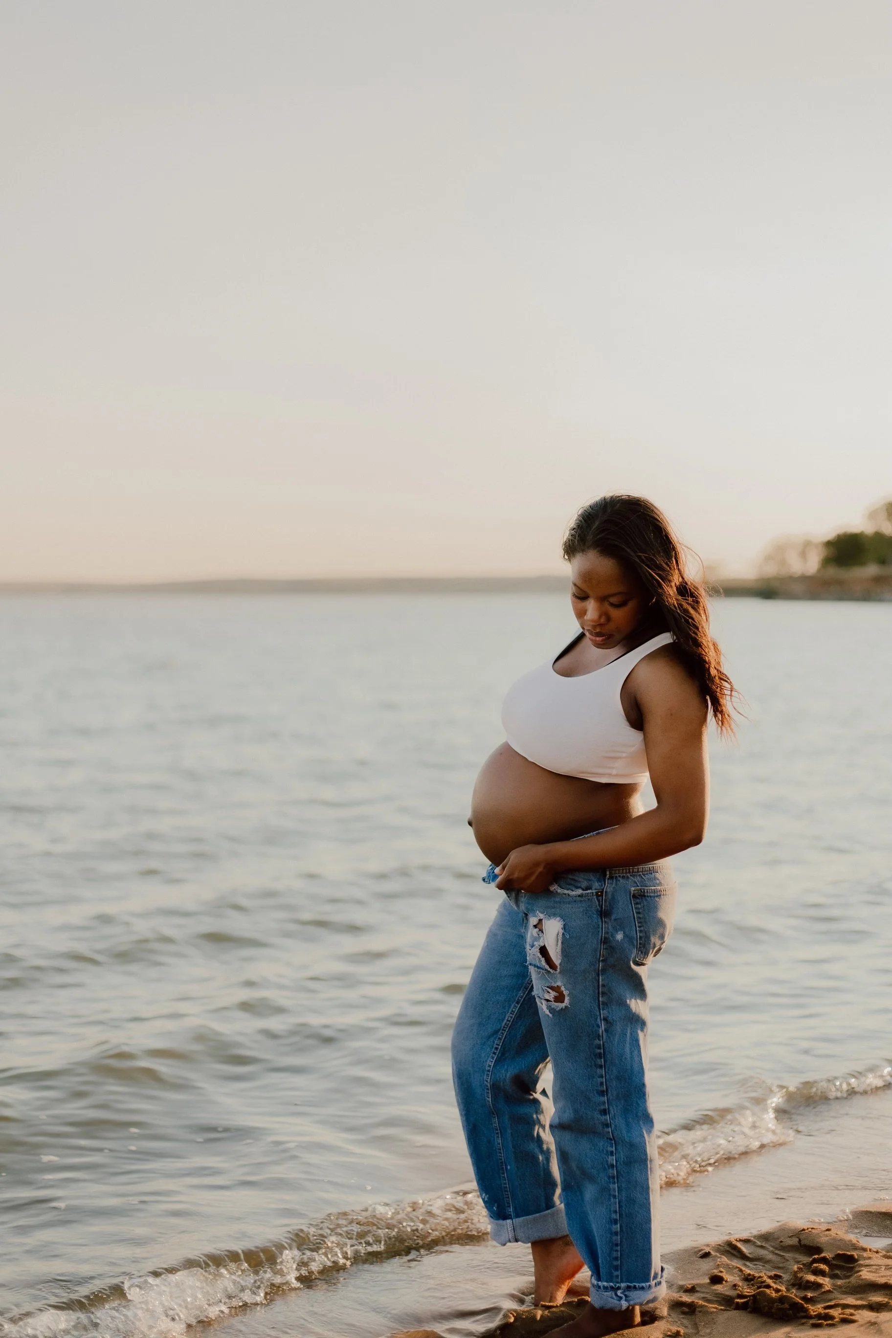 A pregnant woman standing in shallow water at the beach during sunset, wearing a white tank top and ripped jeans.