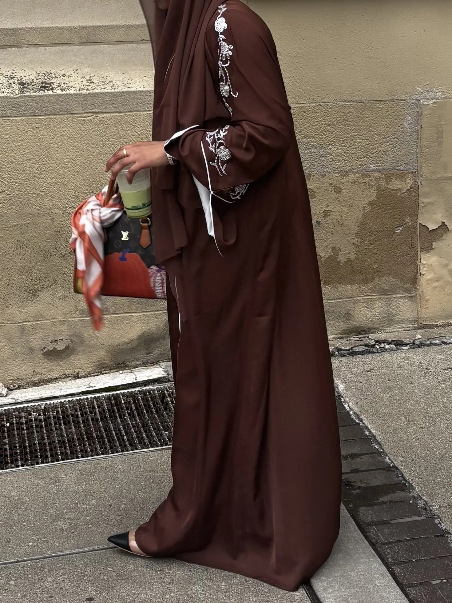 Person dressed in a long, dark brown embroidered dress holding a drink and a Louis Vuitton handbag with a red and white scarf, standing on a concrete sidewalk next to a beige stone wall.