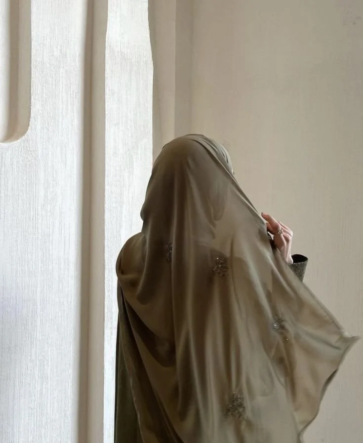 A woman wearing a beige headscarf with floral embroidery, partially covering her face, standing indoors near a wall.