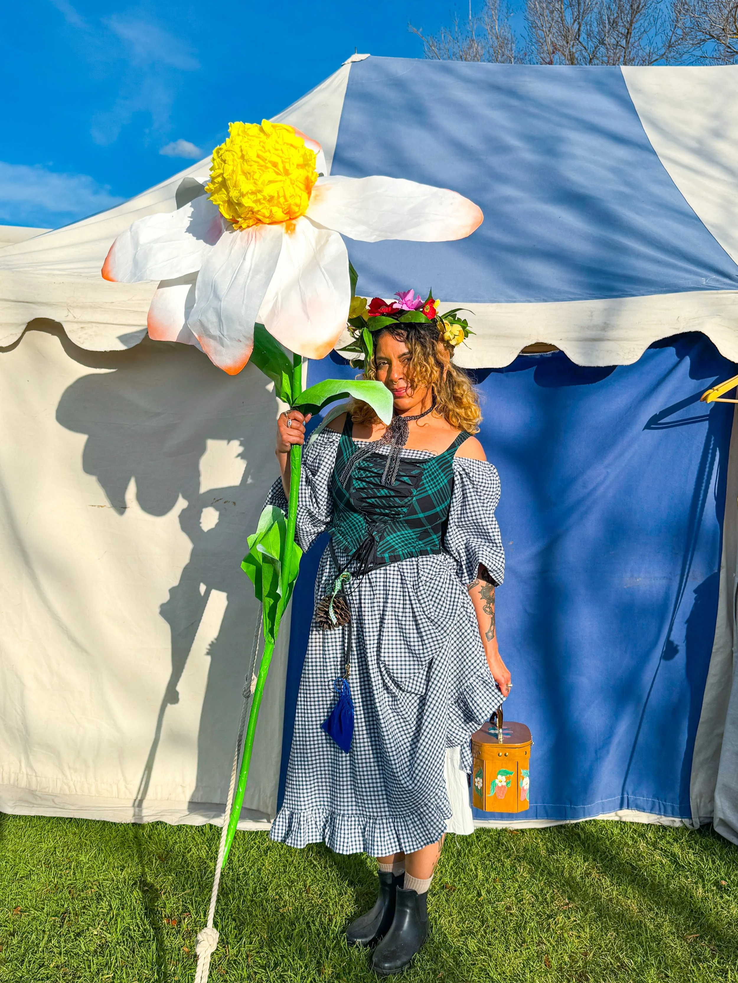 A woman dressed as an elf stands in front of a tent holding a giant papercraft daisy prop