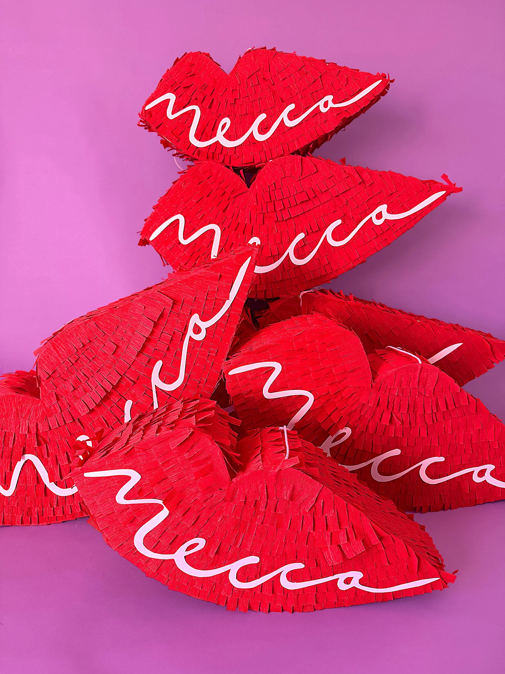 A collection of red lip-shaped piñatas handmade by Kitiya Palaskas are piled on a purple background. Each piñata has a pink Mecca logo on it. 