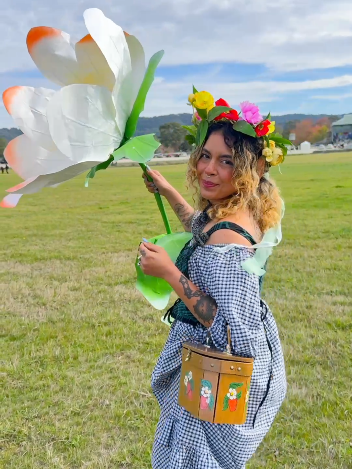 A woman dressed as an elf carries a giant daisy prop through a field, smiling.