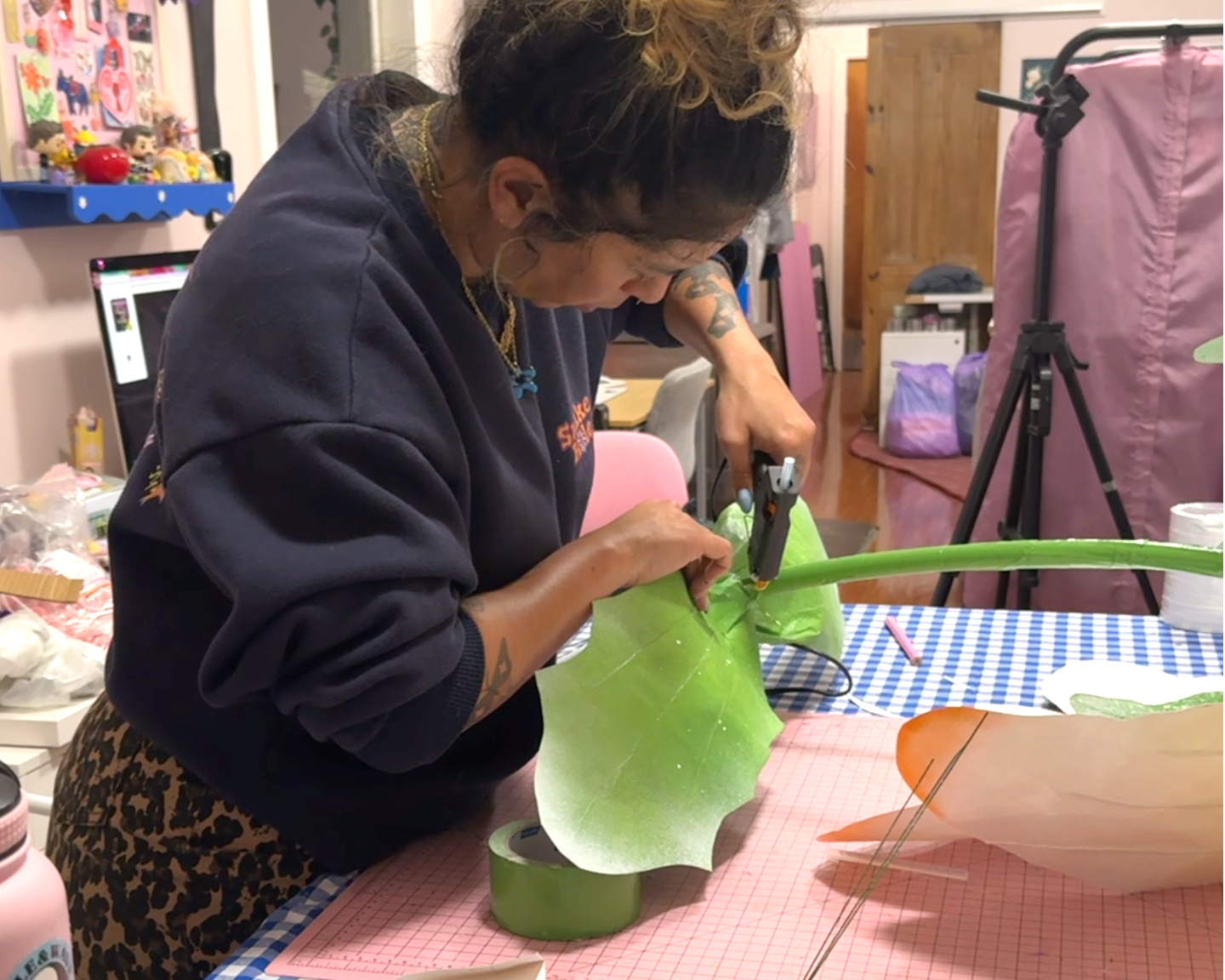 A woman assembles the stem of a giant daisy prop in a messy studio