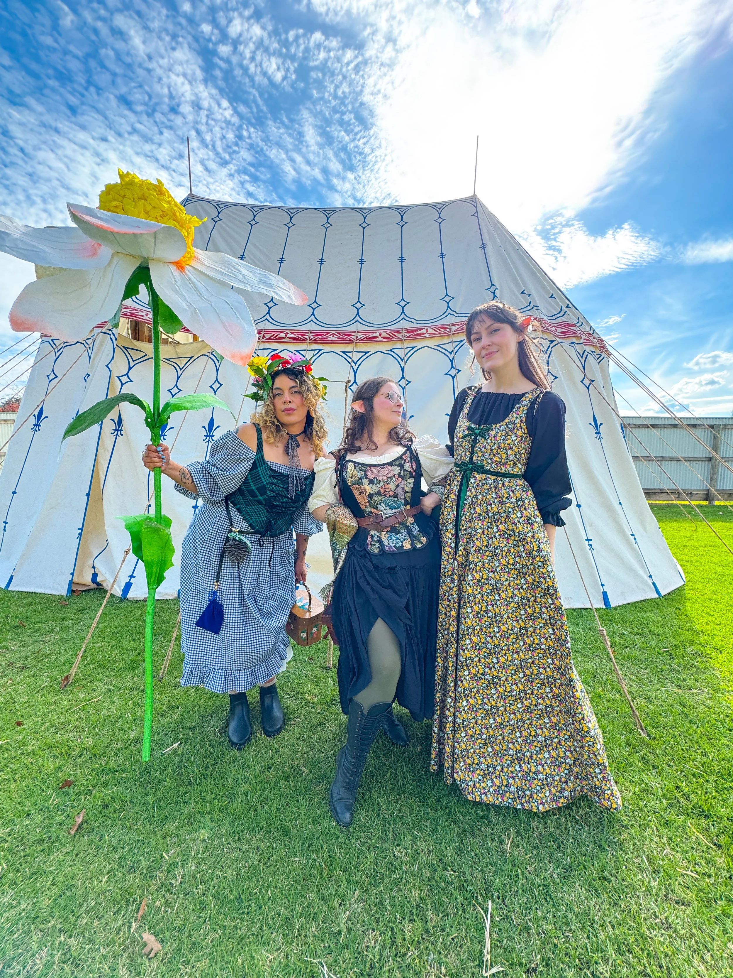 3 women dressed in renaissance costumes stand in front of a whimsical canvas tent. One woman holds a giant papercraft daisy prop.