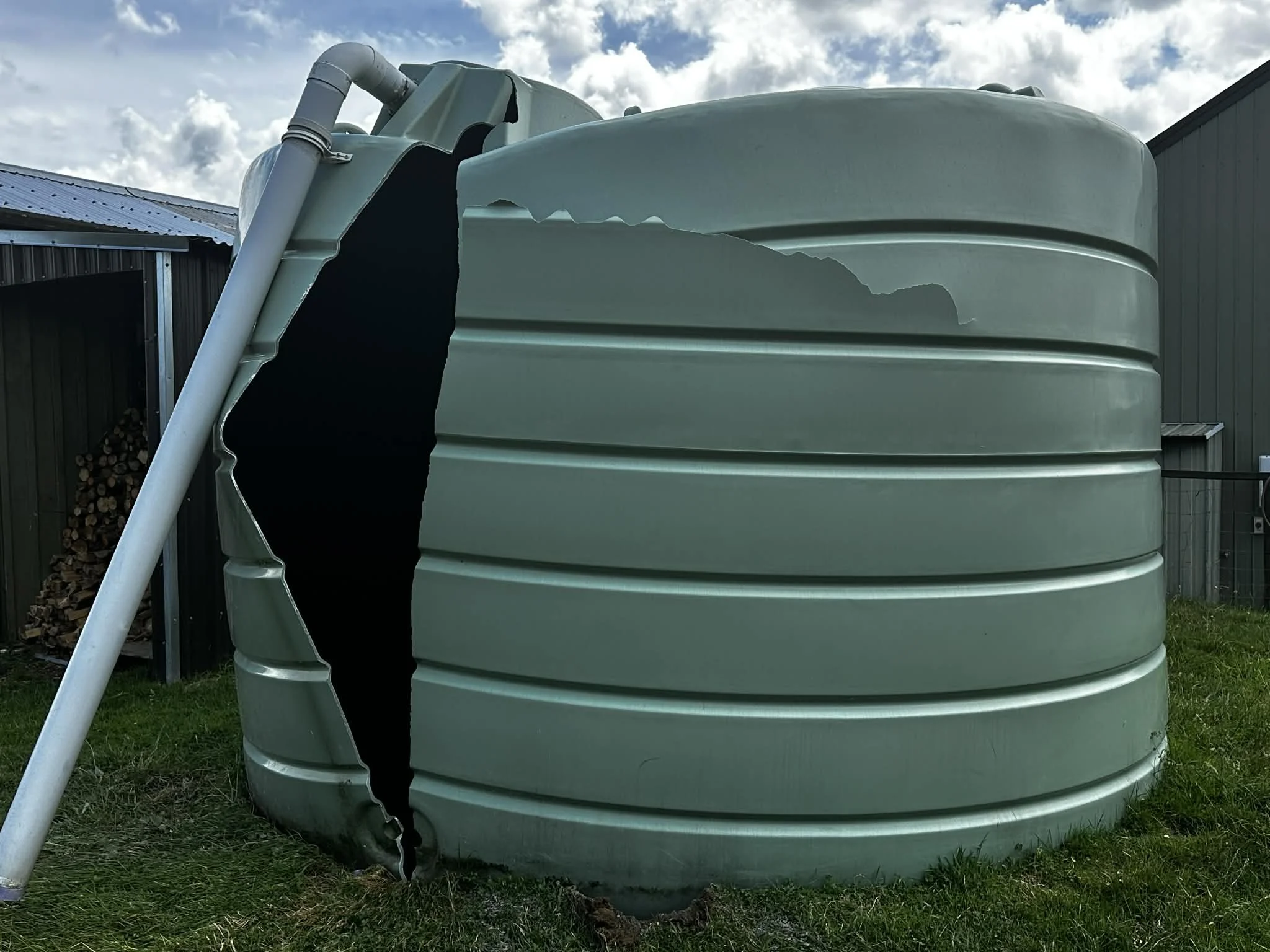 Green metal water storage tank with a large tear, located outdoors on grass, with buildings and a cloudy sky in the background.