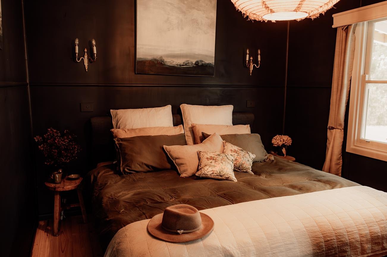 A cozy bedroom with dark walls, a large bed with multiple pillows, a hat on the foot of the bed, side tables with flowers, patterned pillows, wall sconces, a window with curtains, and a painting above the headboard.