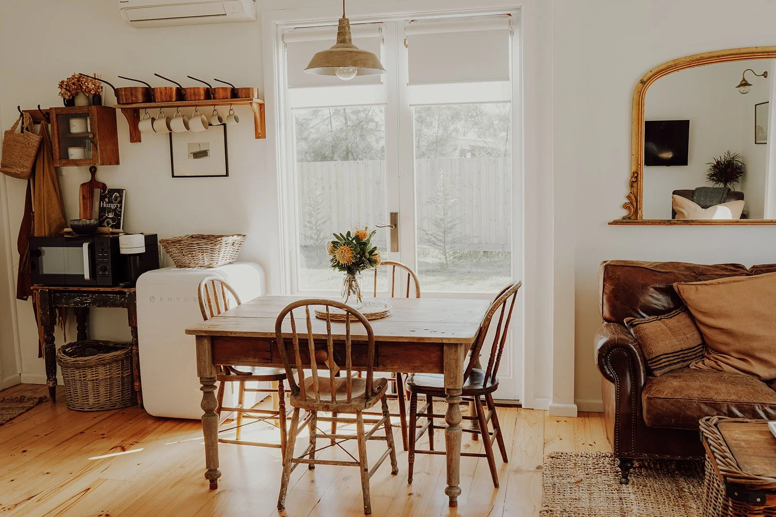 Cozy dining room with a wooden table and chairs, a sliding glass door with white blinds, a mirror on the wall, a brown sofa with pillows, and vintage decor.