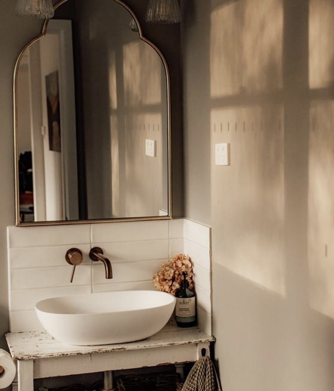 A bathroom vanity with a white oval vessel sink, copper faucet, and a large mirror reflecting a hallway. A bottle of hand soap and a pink floral arrangement sit on the vanity, which is distressed white wood.