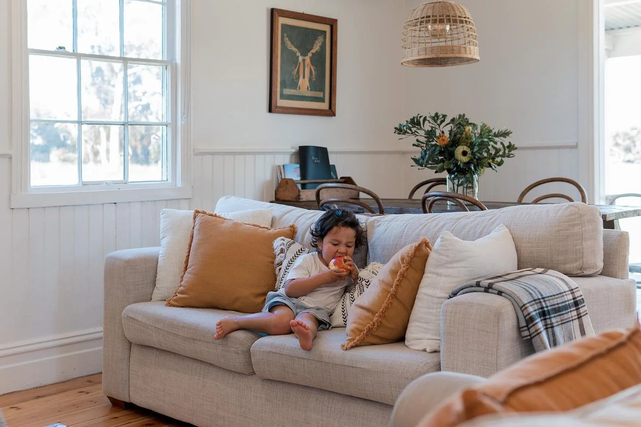 A young child with curly hair sitting on a beige sofa, holding and eating an apple, in a bright living room with white walls, a window, and decorative pillows.