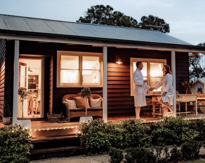 Two people in robes exchanging a handshake on a wooden porch of a house at dusk, with interior lights shining through windows and decorative string lights on the porch