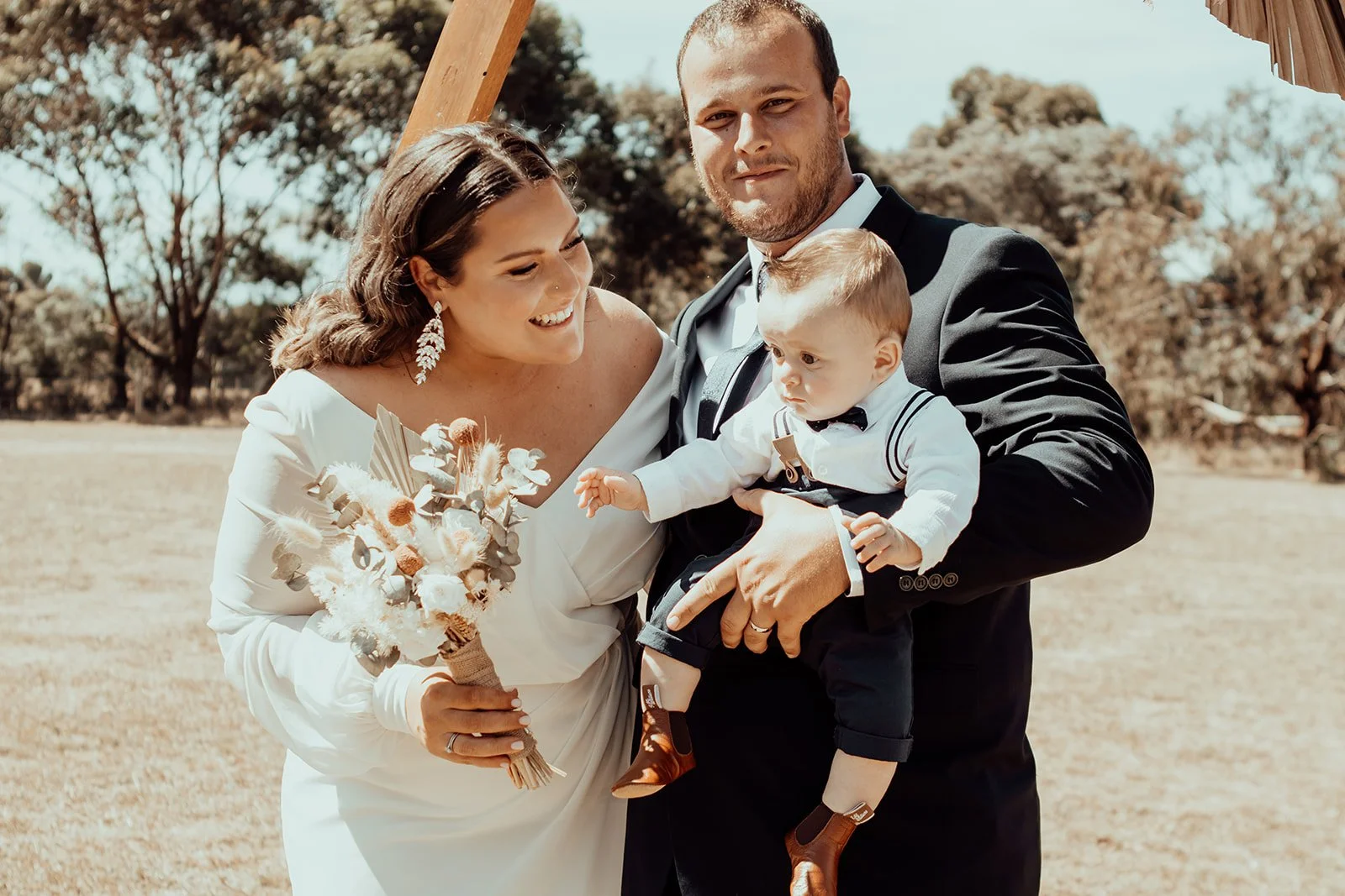 A woman in a white wedding dress holding a bouquet of flowers, smiling and leaning towards a man in a black suit holding a young boy dressed formally with a bow tie. They are outdoors with trees in the background.