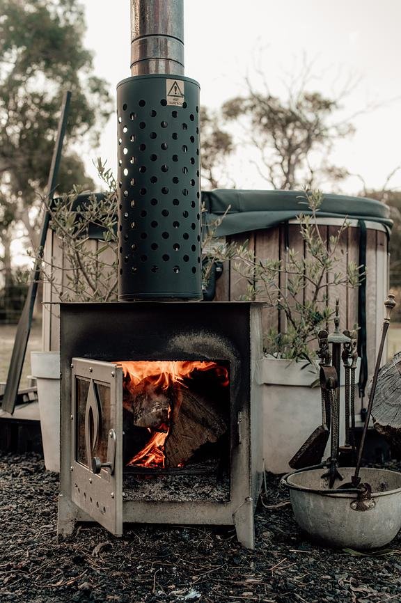 A wood-burning outdoor stove with a fire inside, connected to a metal chimney pipe, surrounded by gardening plants and tools.