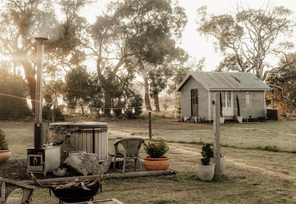 A rural backyard scene during sunset with a small house, potted plants, a wood stove, and trees in the background.