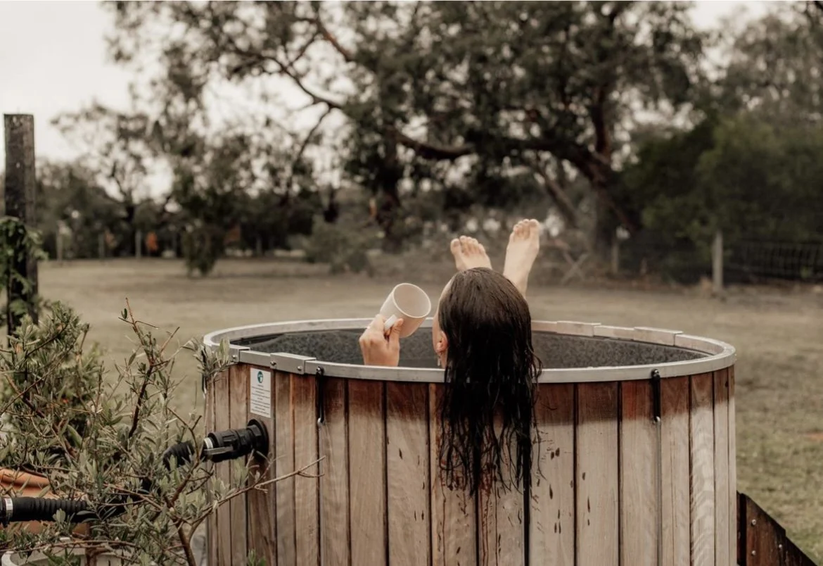 Person with long wet hair relaxing in a wooden hot tub outdoors, holding a cup and with legs up, surrounded by trees and a fence.