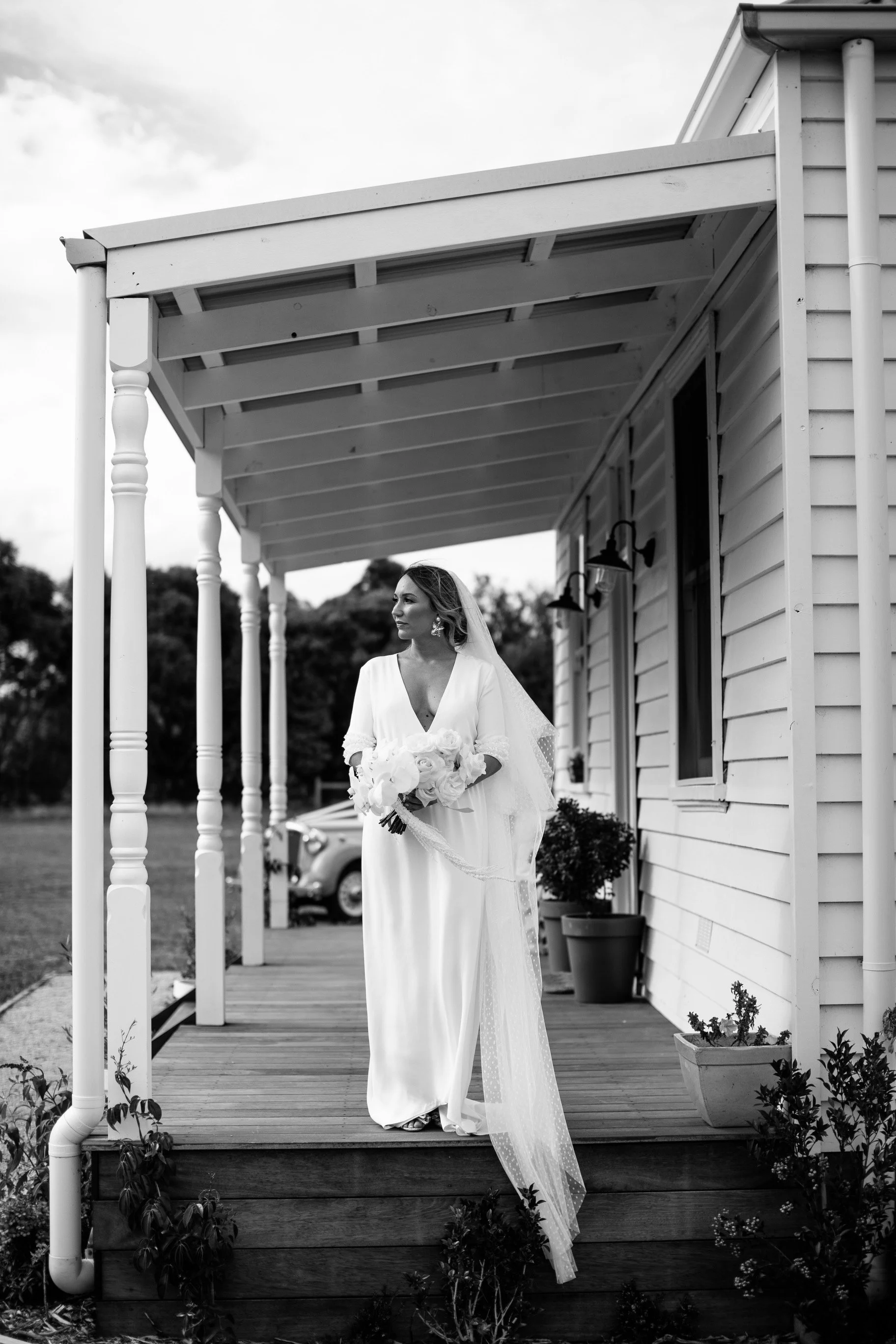 A bride in a white dress holding a bouquet of flowers standing on a porch of a house with white siding, outdoor lighting, and flower pots.