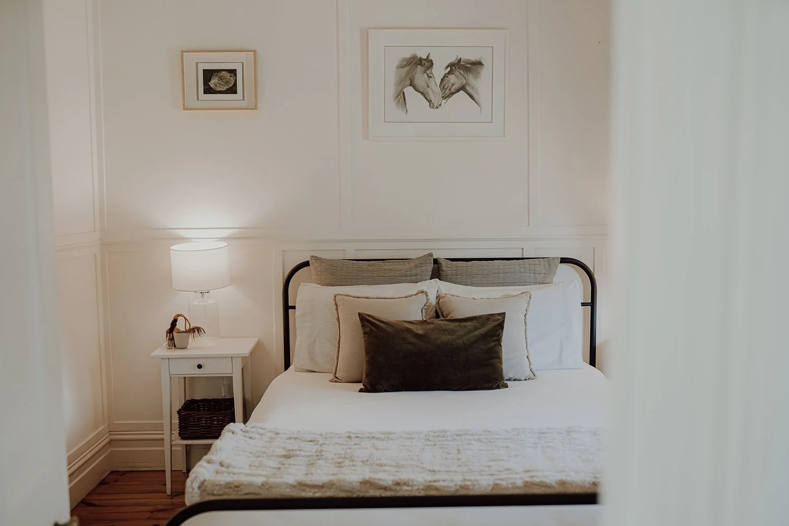 A neatly made bed with white bedding and multiple pillows, positioned against a white wall with framed artwork of two horses and a fish. A small white nightstand with a lamp, a basket, and a decorative item is beside the bed.