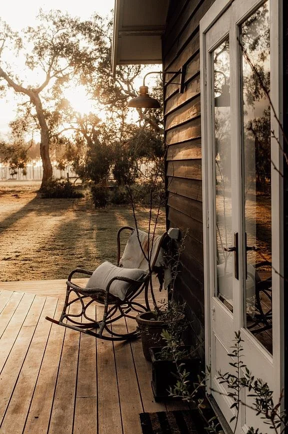 A wooden porch with an antique rocking chair draped with a pillow, potted plants, below a black exterior wall, near a glass door, and trees illuminated by sunset in the background.