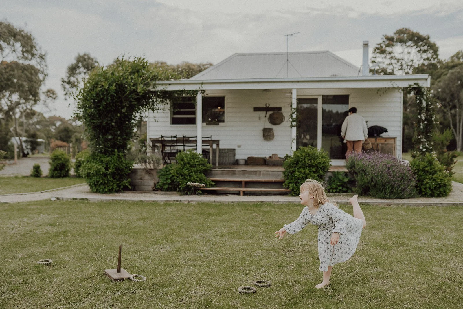 A young girl in a white dress playing in a backyard with a lawn game, in front of a white house with a porch and garden plants.