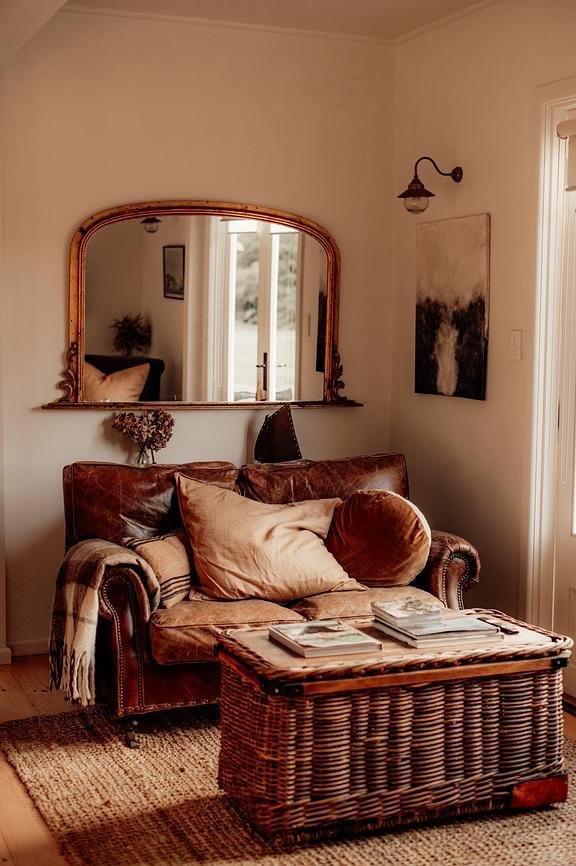 Living room with leather sofa, decorative pillows, a striped blanket, a wicker coffee table with magazines, a large mirror on the wall, a framed artwork, wall-mounted lamp, and a sliding glass door.