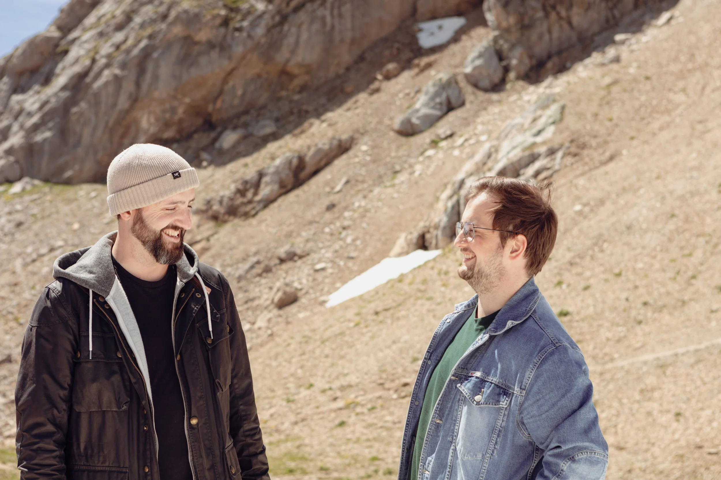 Ben Hodson and Benji Amman standing outdoors on a mountain with rocky terrain in the background, smiling and engaging in conversation. One wears a beanie and a black jacket, the other has glasses and a denim jacket.