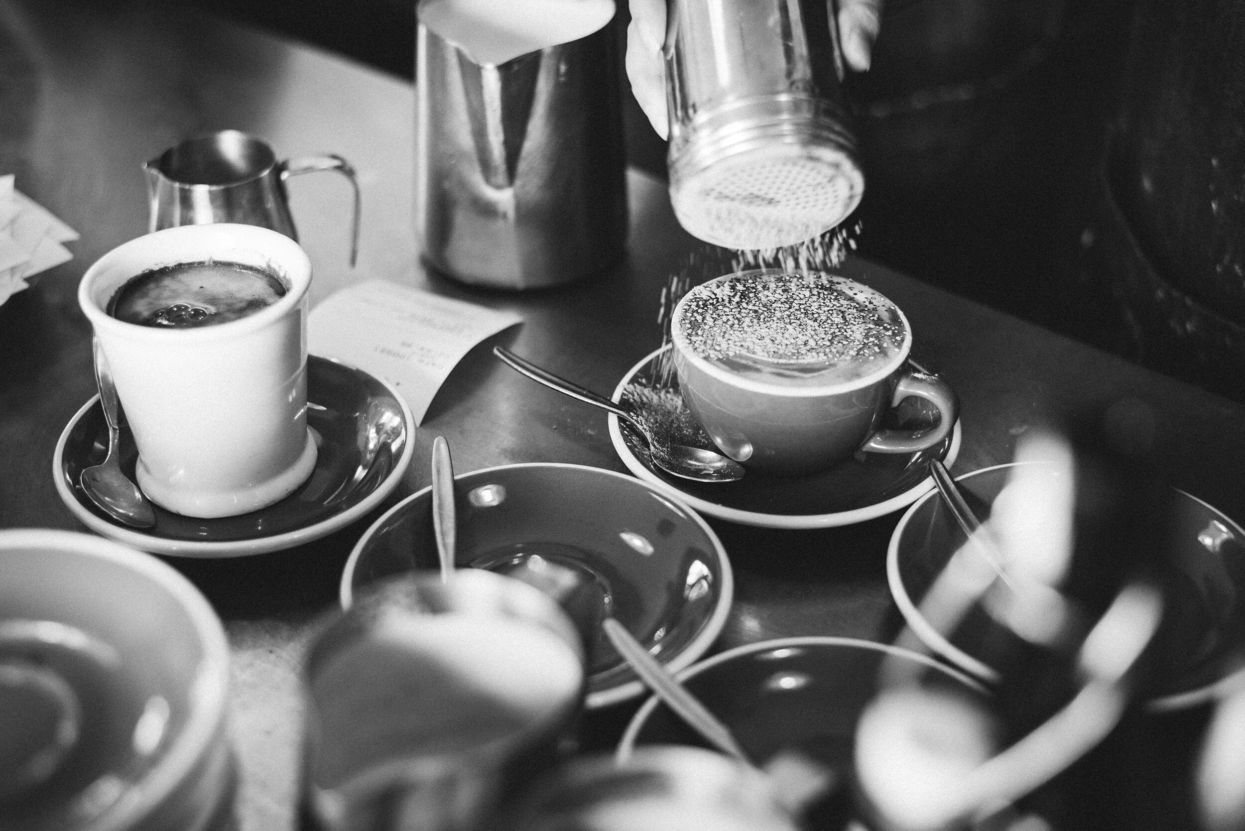 Black and white photo of coffee cups, some with saucers, on a table with a coffee pot pouring sugar into a cup, surrounded by spoons and small bowls.