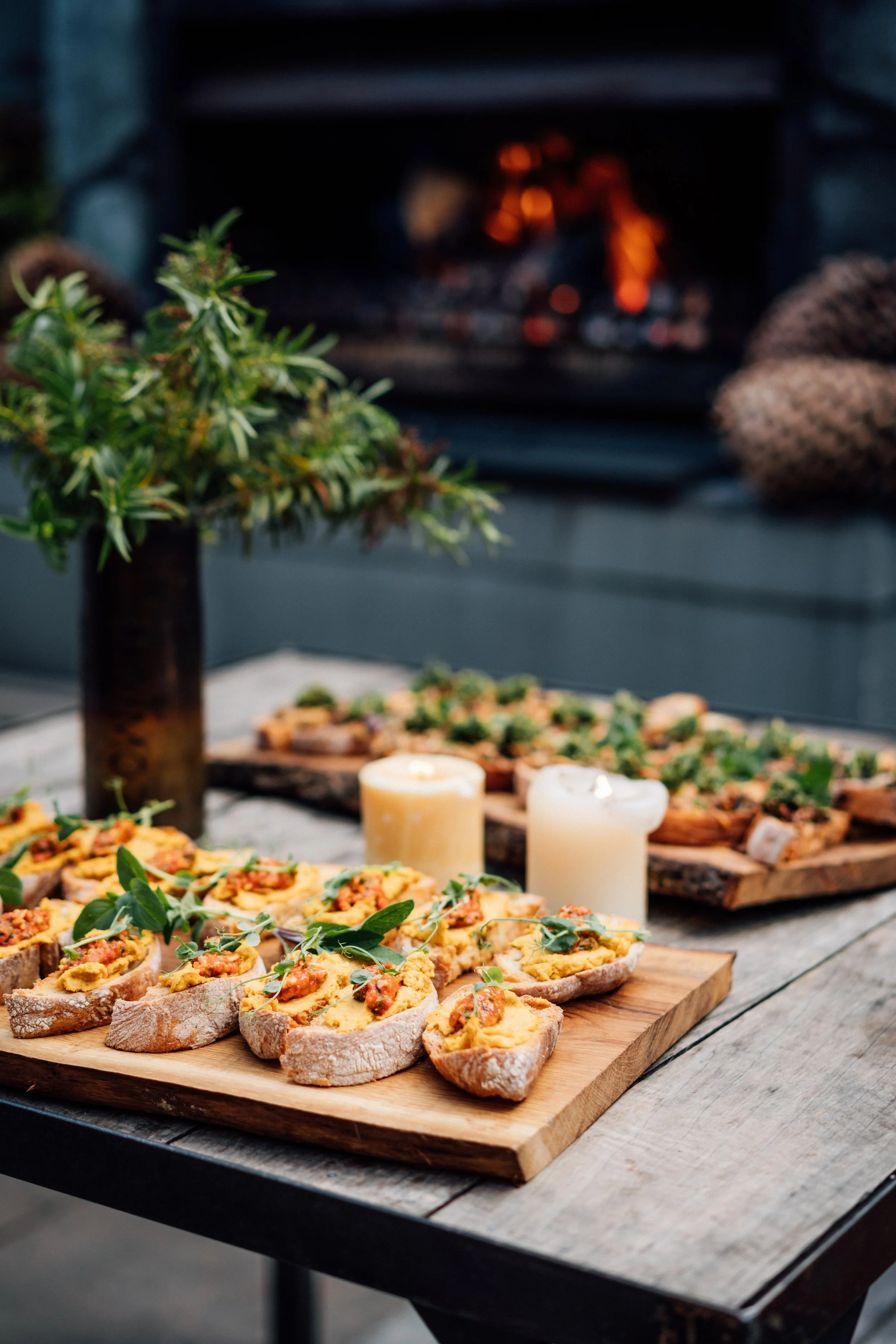 Arranged food on a wooden tray with small pieces of bread topped with egg salad and garnished with herbs. A tall vase with green foliage and two lit candles are on the table. In the background, a fireplace with a fire burning and a knitted item are v