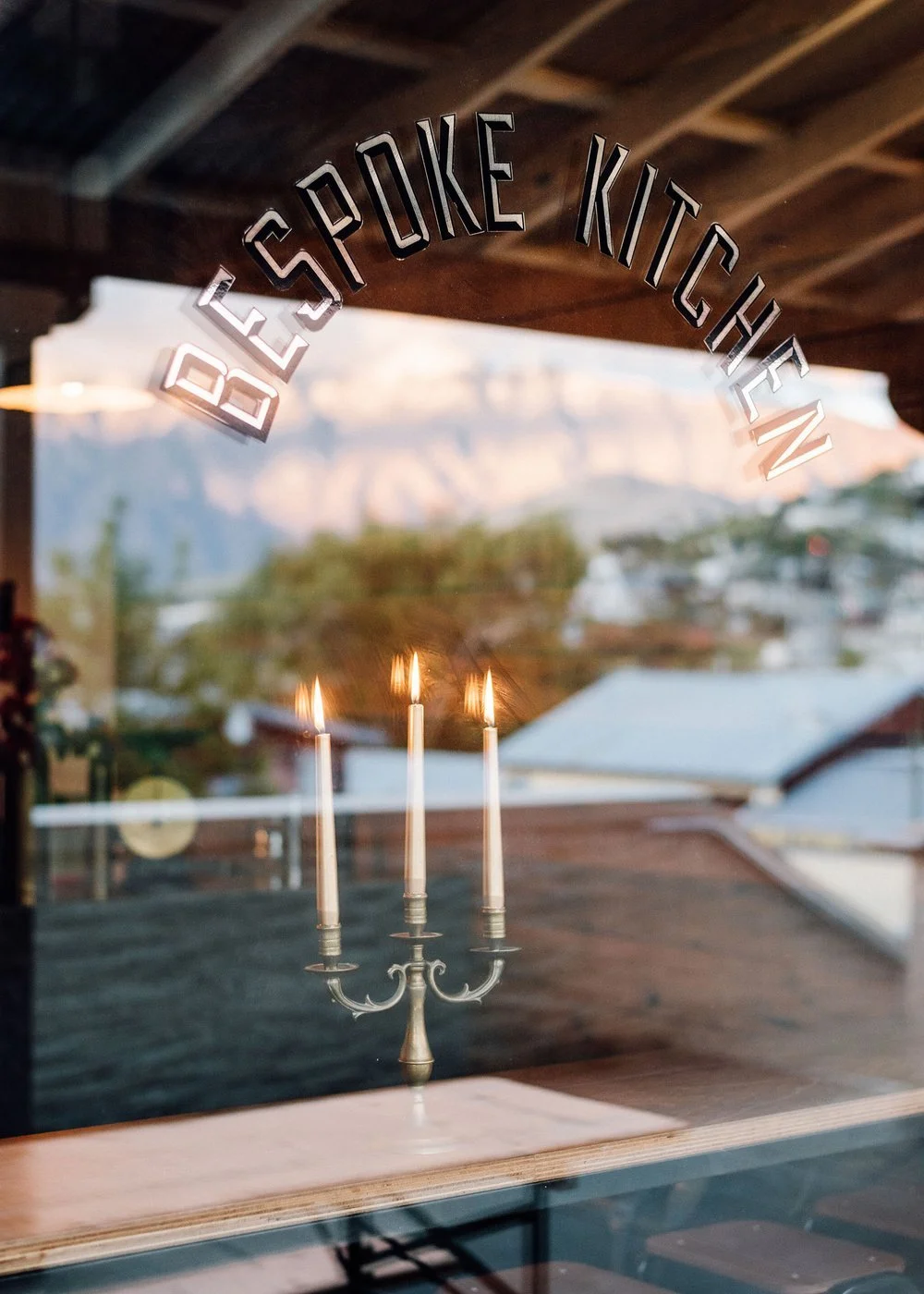 Glass window with the words "Be Spoke Kitchen" written on it, showing reflections of three lit candles and a mountain landscape outside.