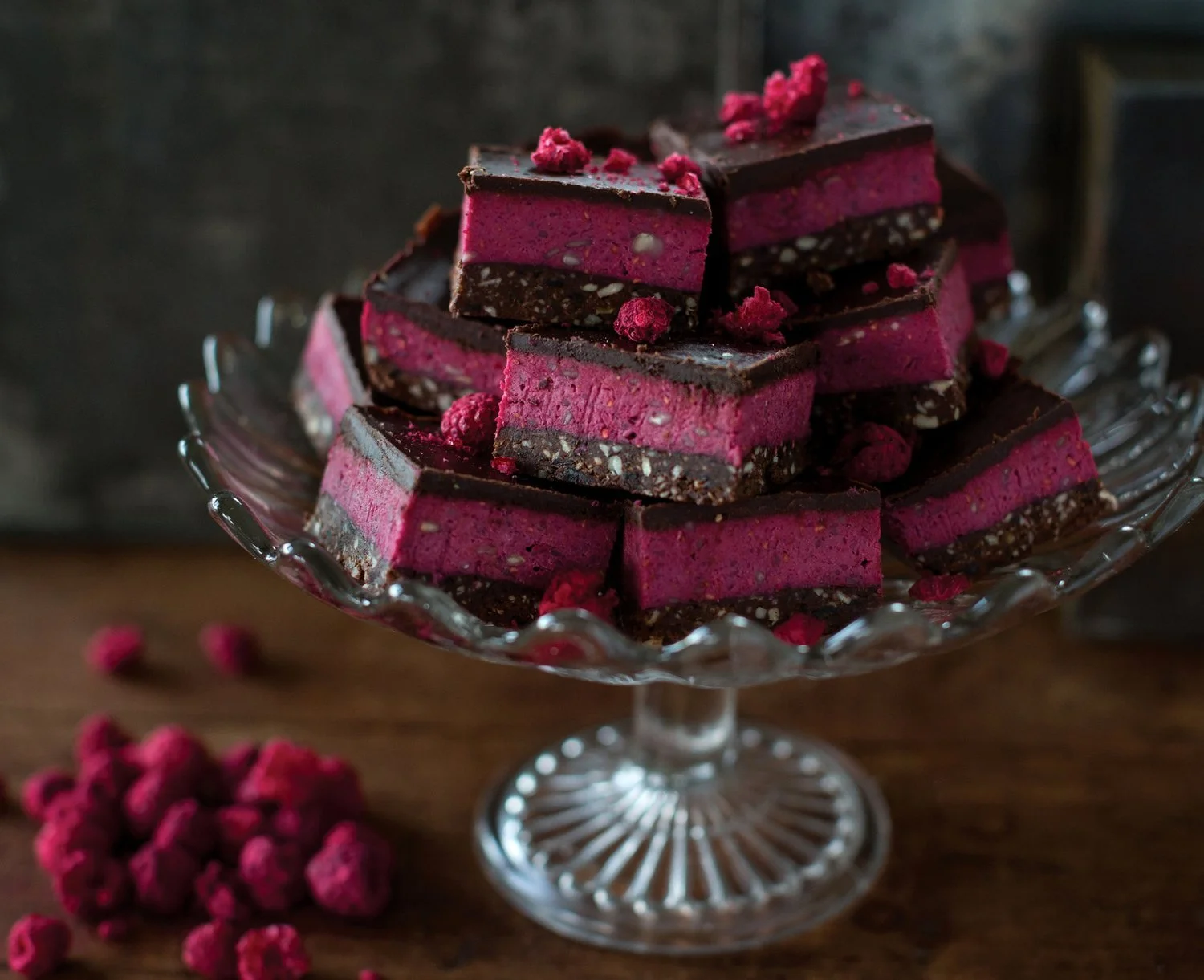 Stacked pink and chocolate layered dessert squares on a glass cake stand with a few berries on a wooden surface.