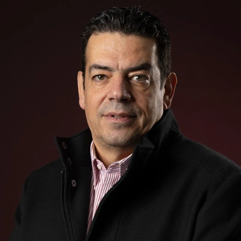 A close-up portrait of a man with dark hair, wearing a black jacket and a pink-striped button-down shirt, against a dark red background.